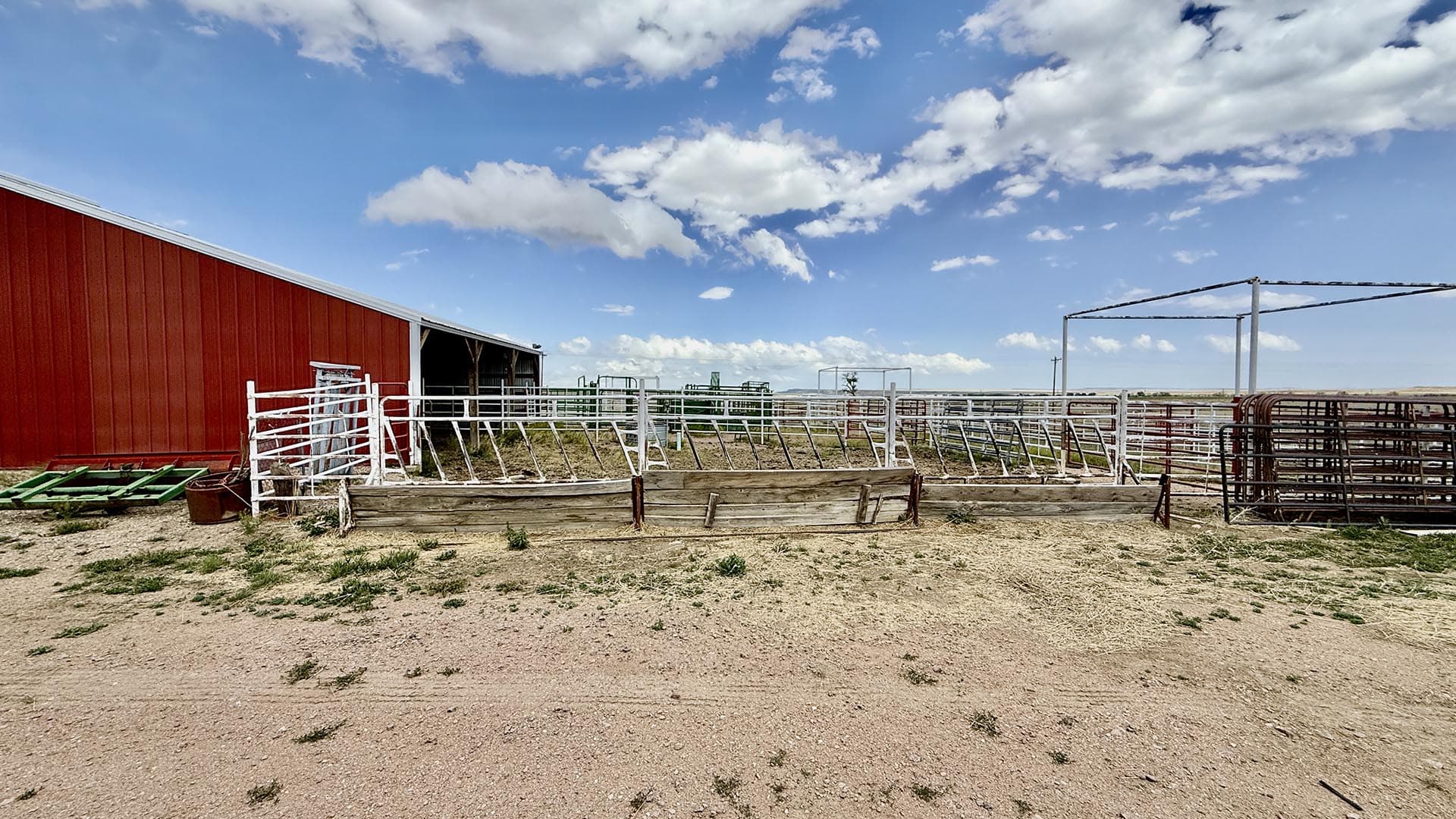 Red barn with white metal fencing and cattle pens in a dry farmyard under a blue sky with scattered clouds