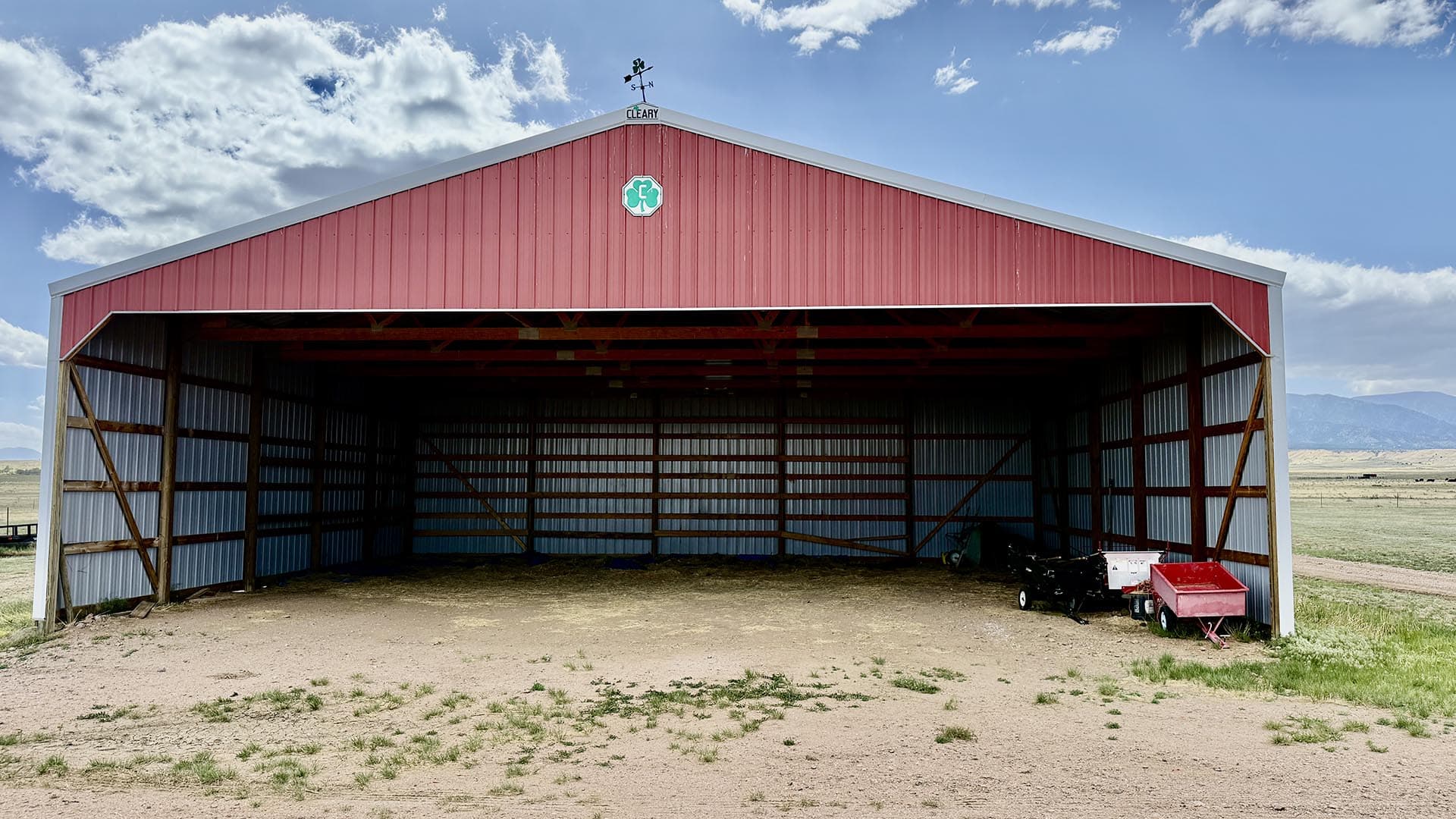 Open red-roofed metal storage barn in a flat rural landscape with farming equipment inside (trailer and red cart).