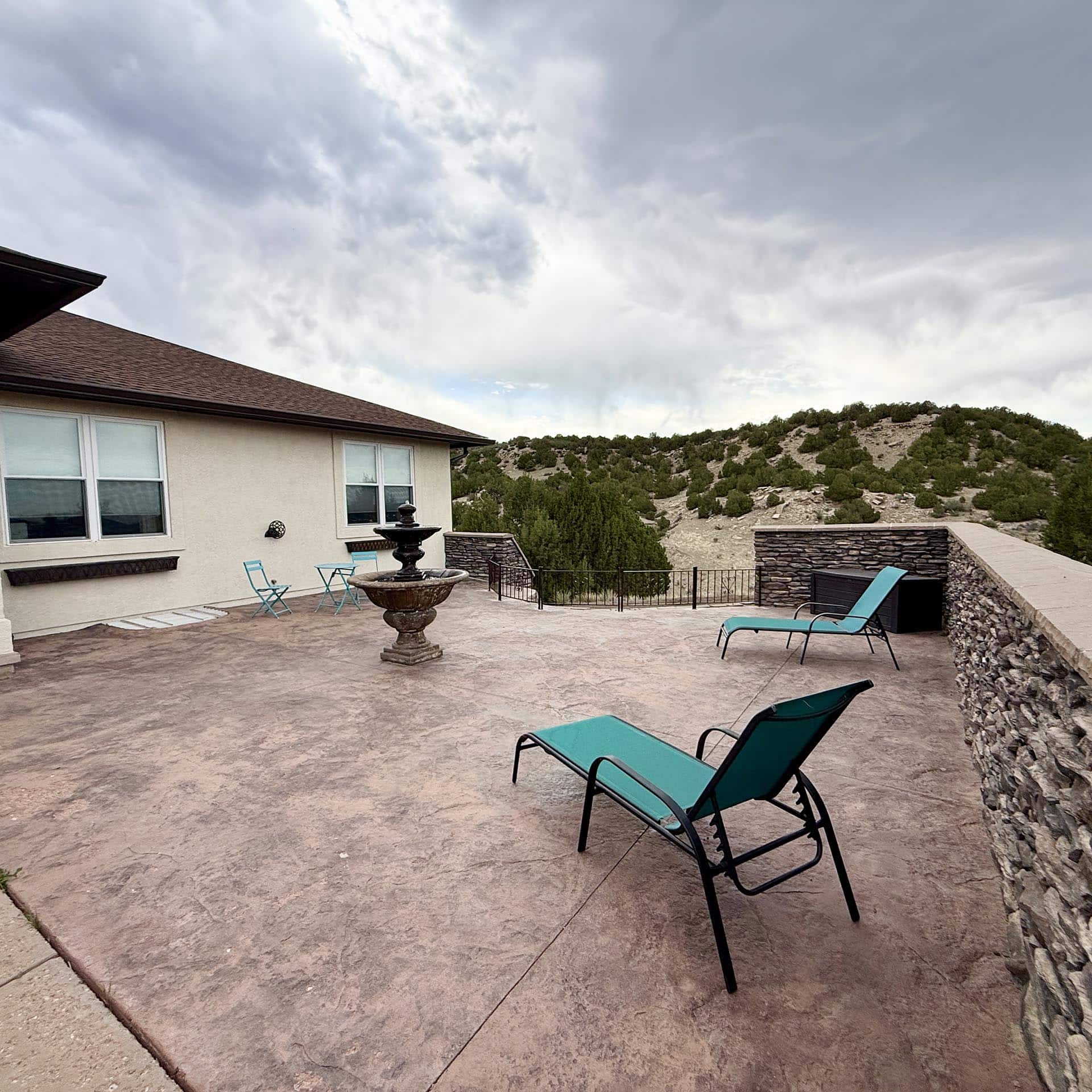 Backyard patio with turquoise chairs around a fountain, overlooking a rocky hillside and cloudy sky.