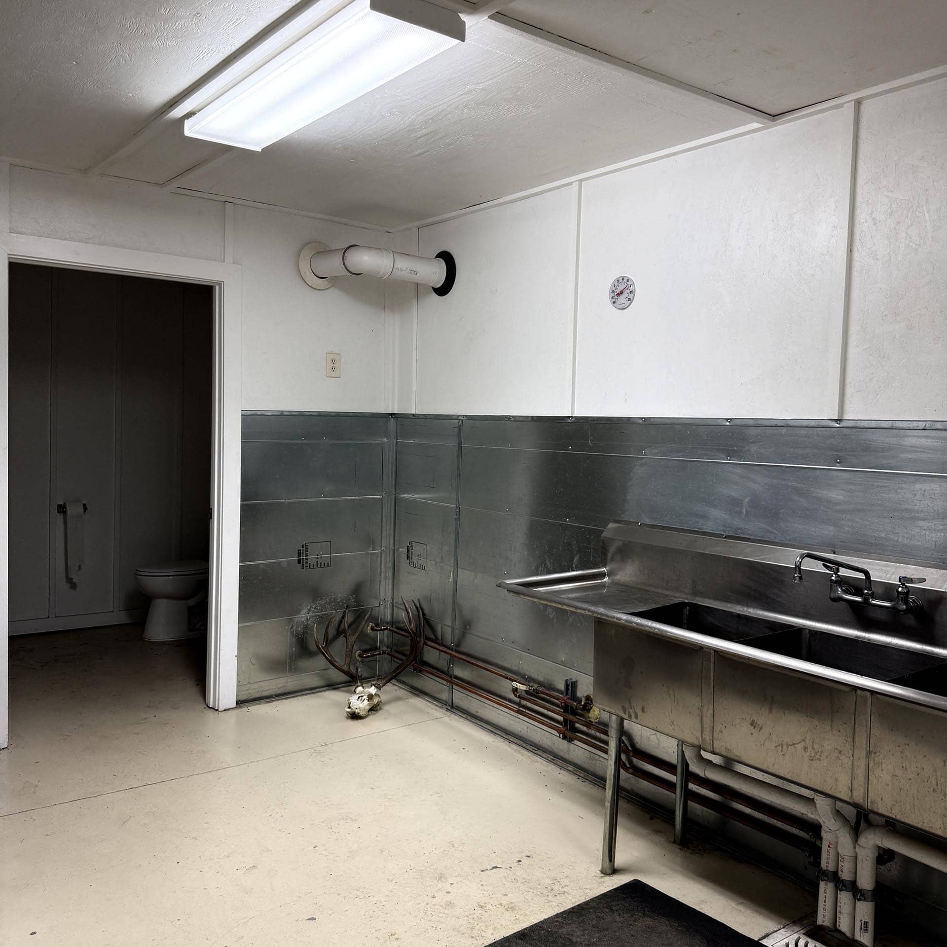 Industrial-style kitchen area with a stainless steel sink and exposed piping along the wall, small thermometer on the white panel, and a doorway revealing a toilet in a dim corner.