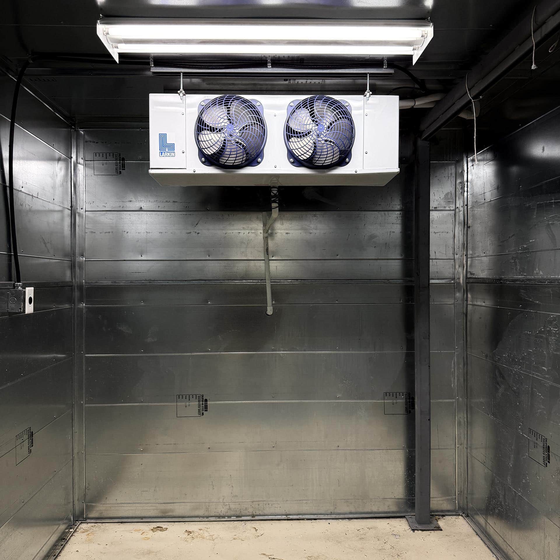 Industrial cooling unit with two blue fans mounted on a white panel, in a stainless steel utility room.