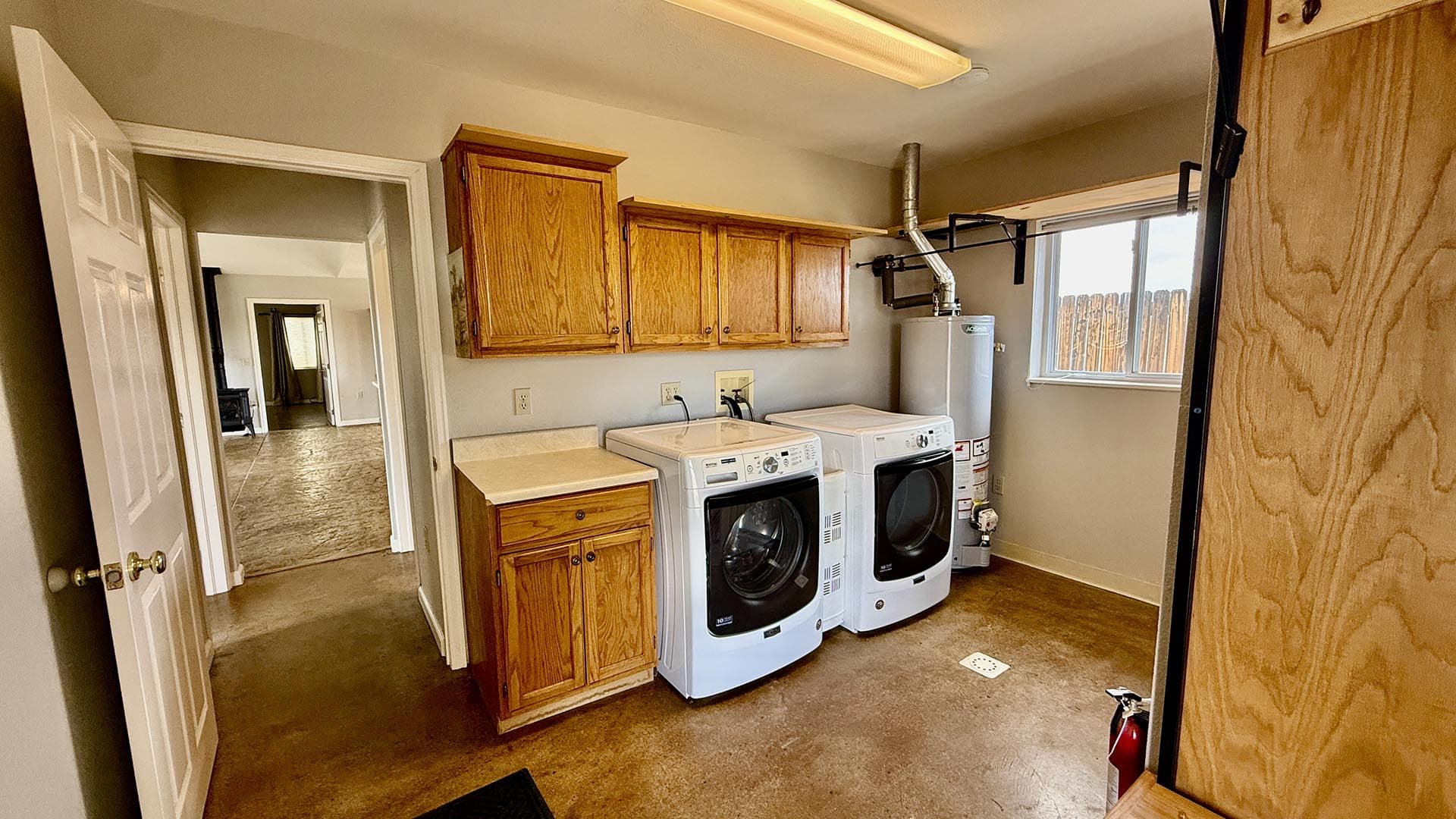 Laundry room with oak cabinets, a washing machine and dryer, and a water heater by a window overlooking a wooden fence outside.