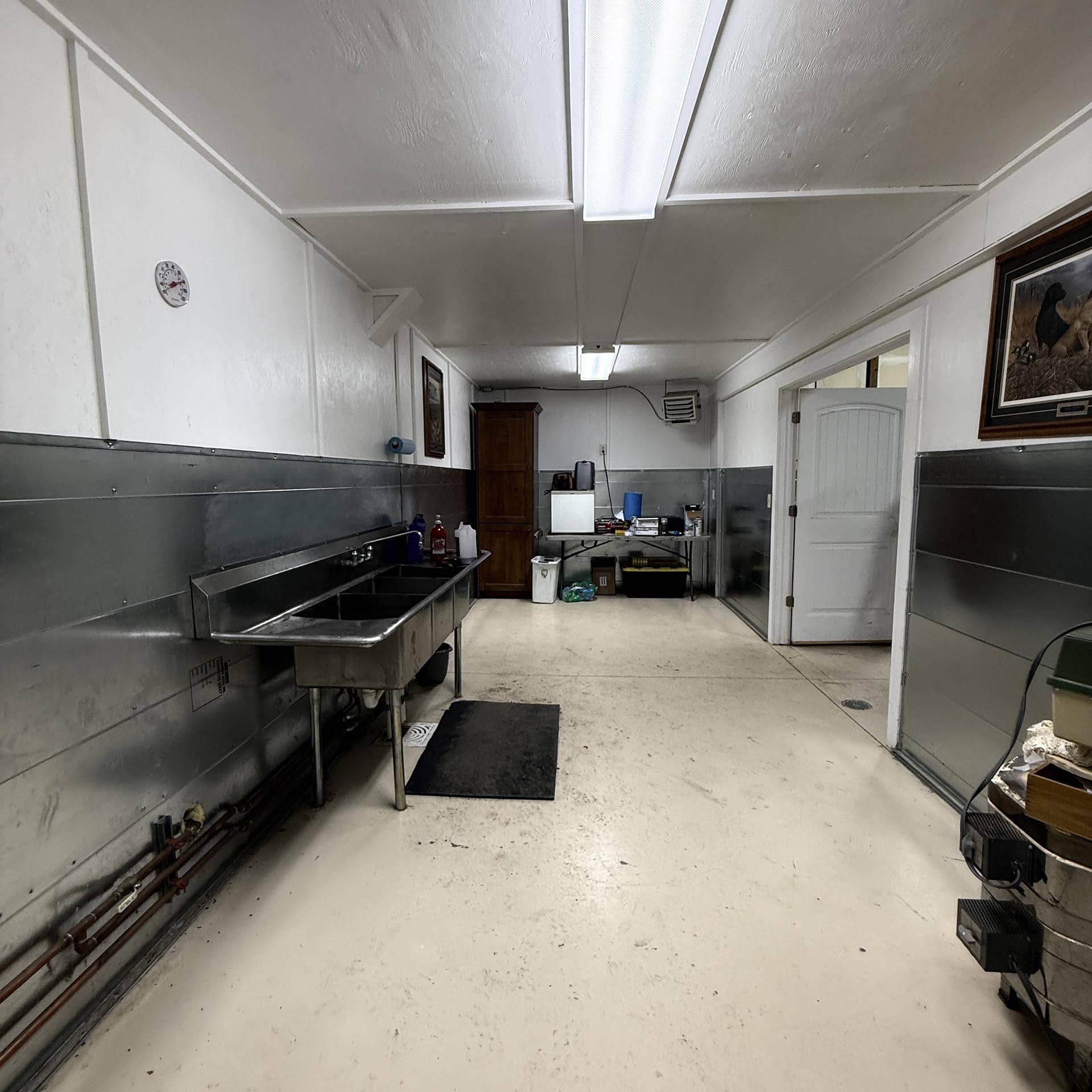 Industrial kitchen with stainless steel sinks along the left wall, white walls, and bright overhead lights; cluttered work area at the far end.