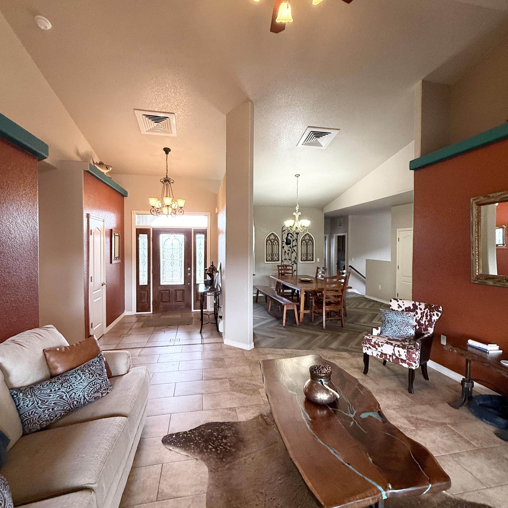 Open-concept living and dining area with red accent walls, beige sofa, patterned throw pillows, and a dark wood coffee table under a chandelier in the dining room.