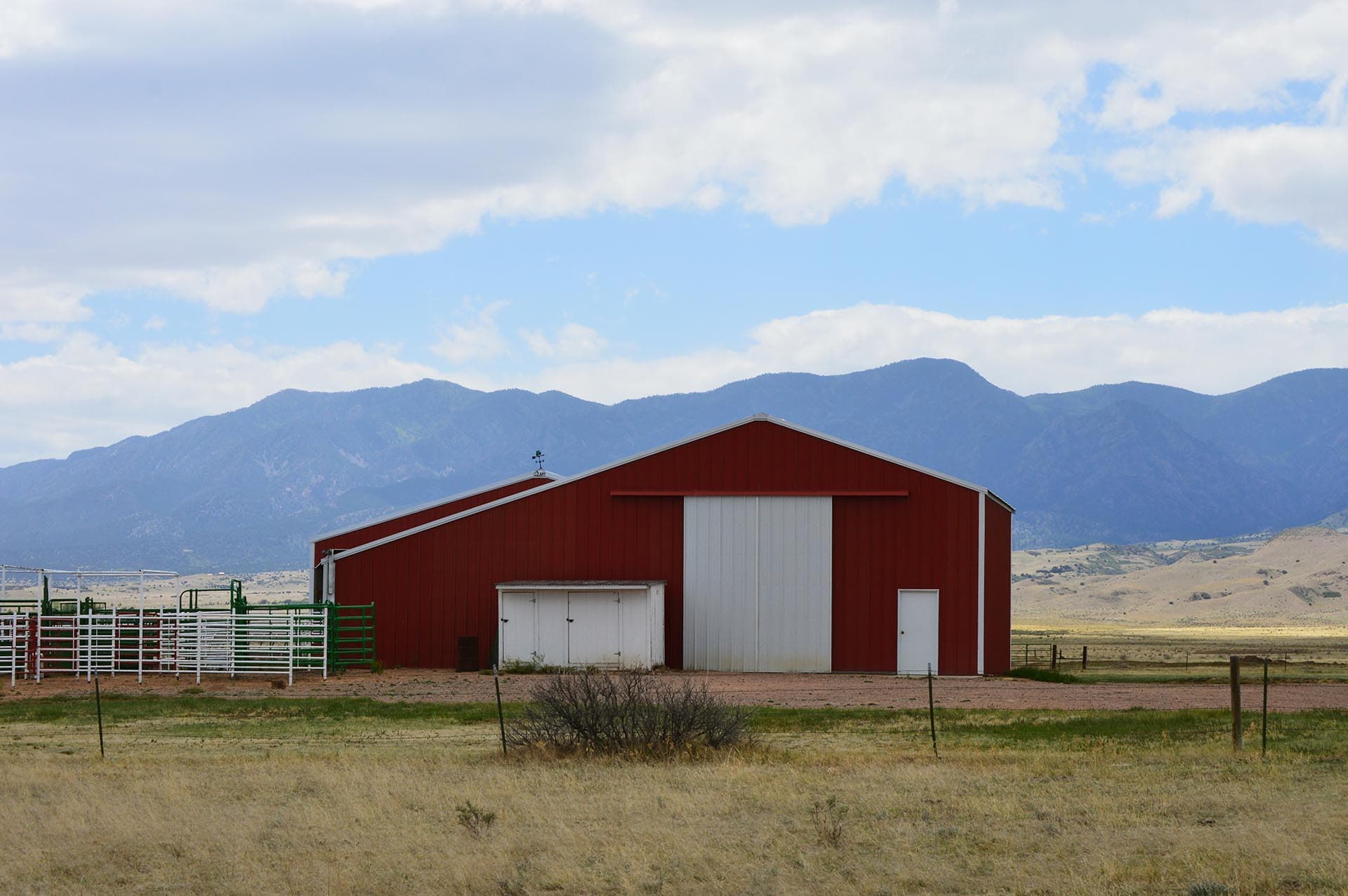 Red barn with white doors sits in a dry field, mountains in the background under a partly cloudy blue sky, a white fence in the foreground.