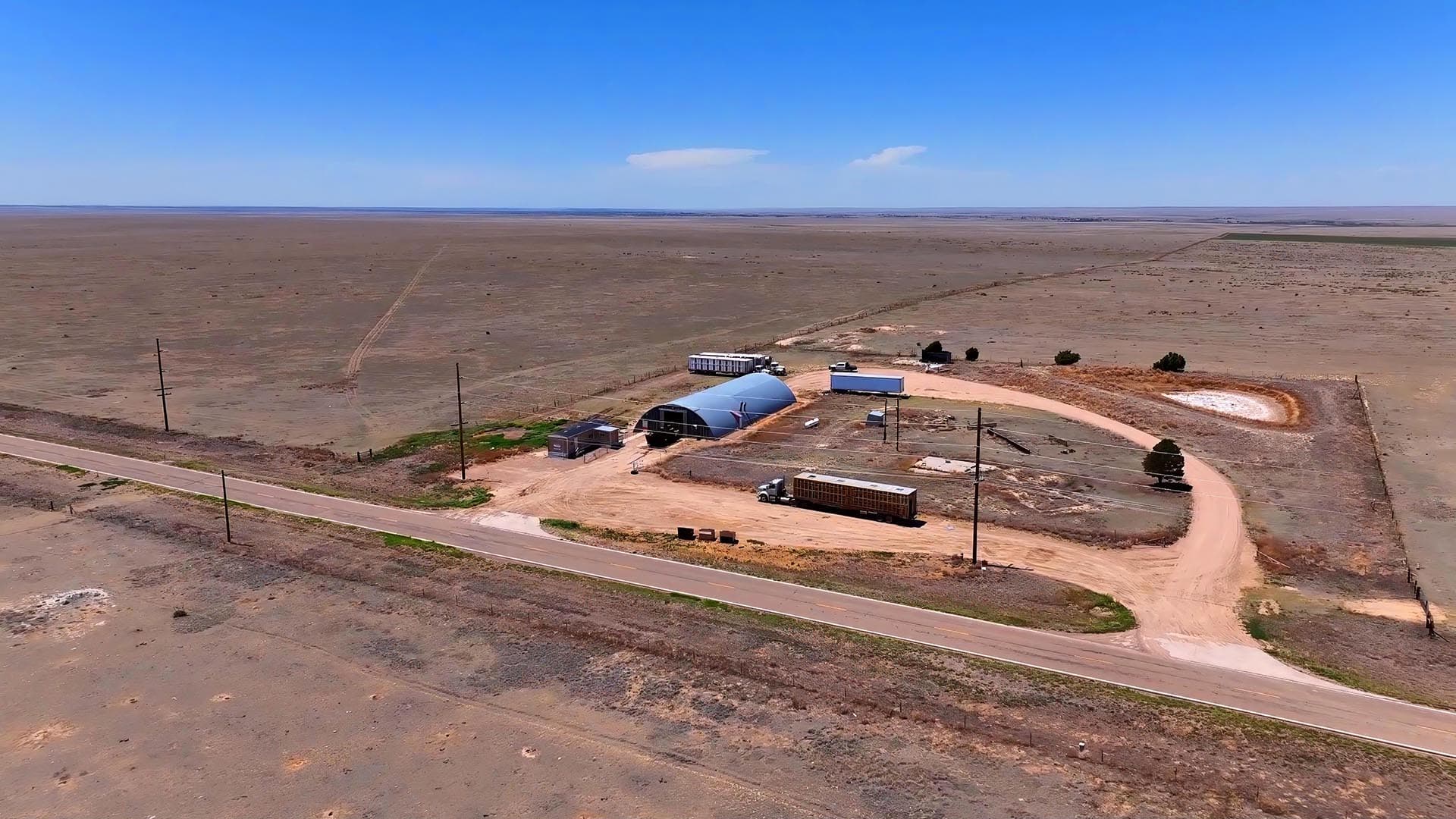 Aerial view of a remote farm complex on a dry, flat plain with a curved dirt road, blue-roofed shelter, trailers, and utility poles.