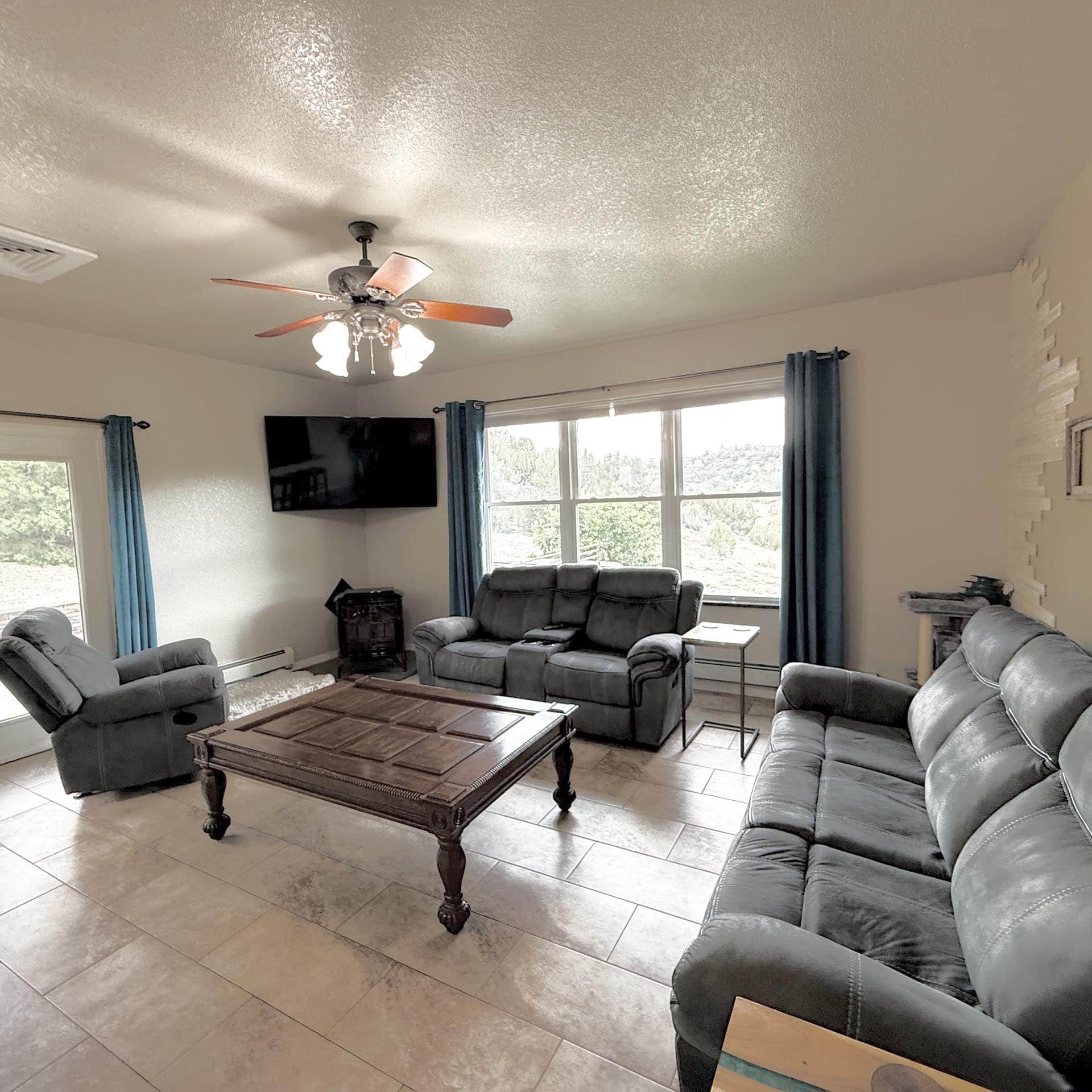 Living room with a ceiling fan, gray leather sofa set, a wooden coffee table, and large windows with blue curtains.