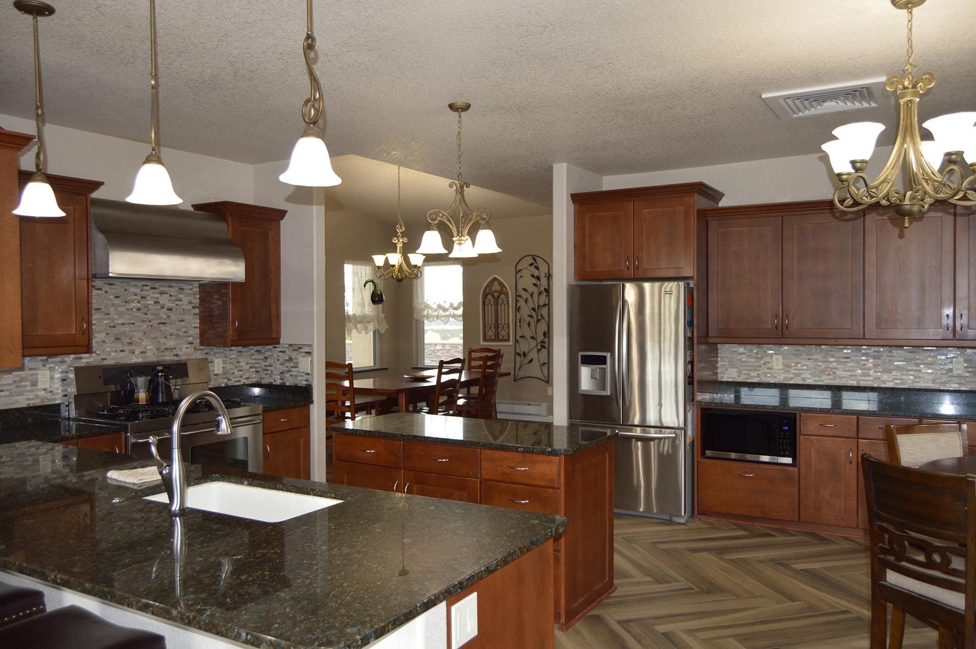 Spacious kitchen with granite island, dark wood cabinets, and stainless appliances, open to a dining nook under decorative chandeliers.