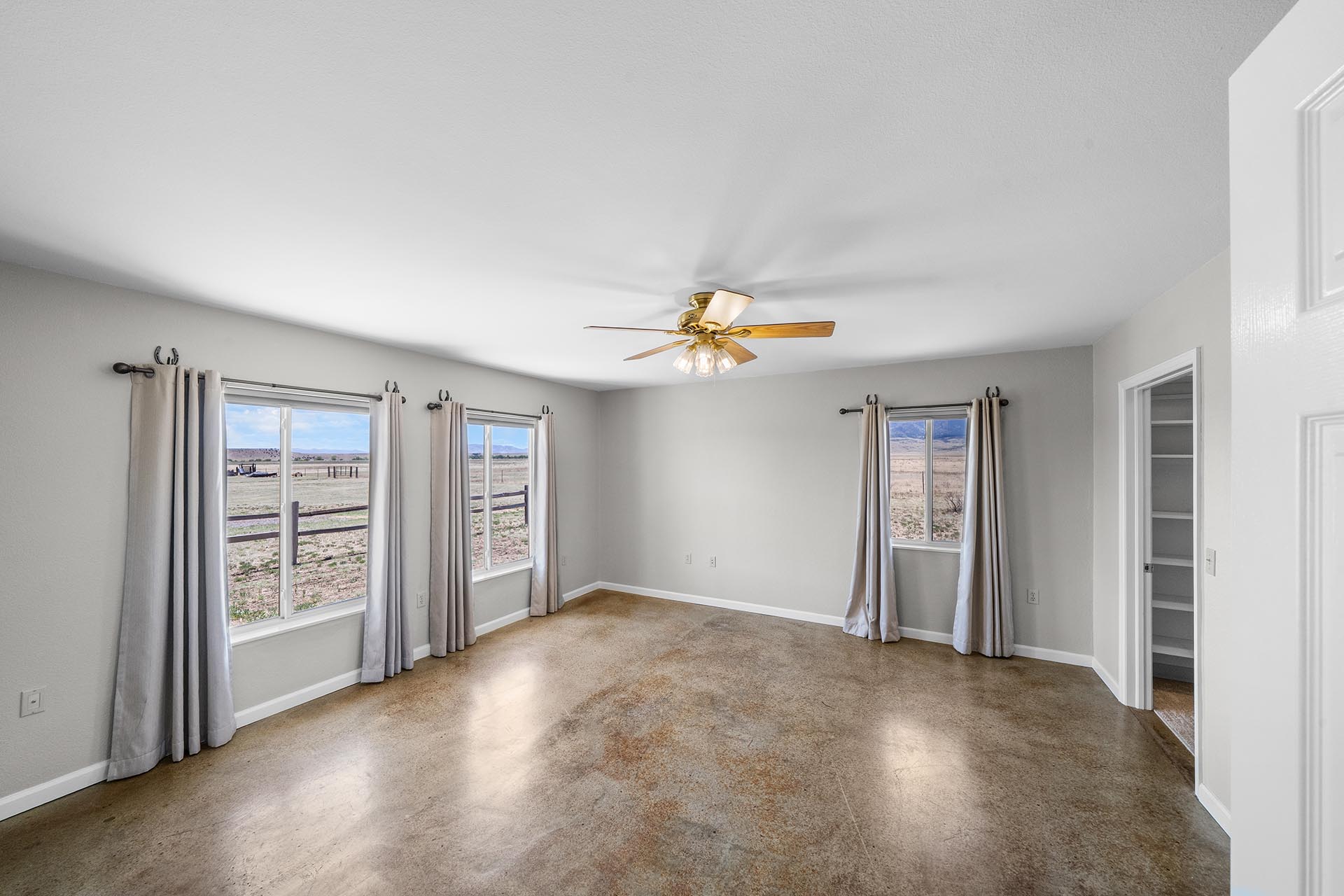 Bright, empty living room with a ceiling fan and large windows overlooking a rural field; neutral walls and carpeted floor