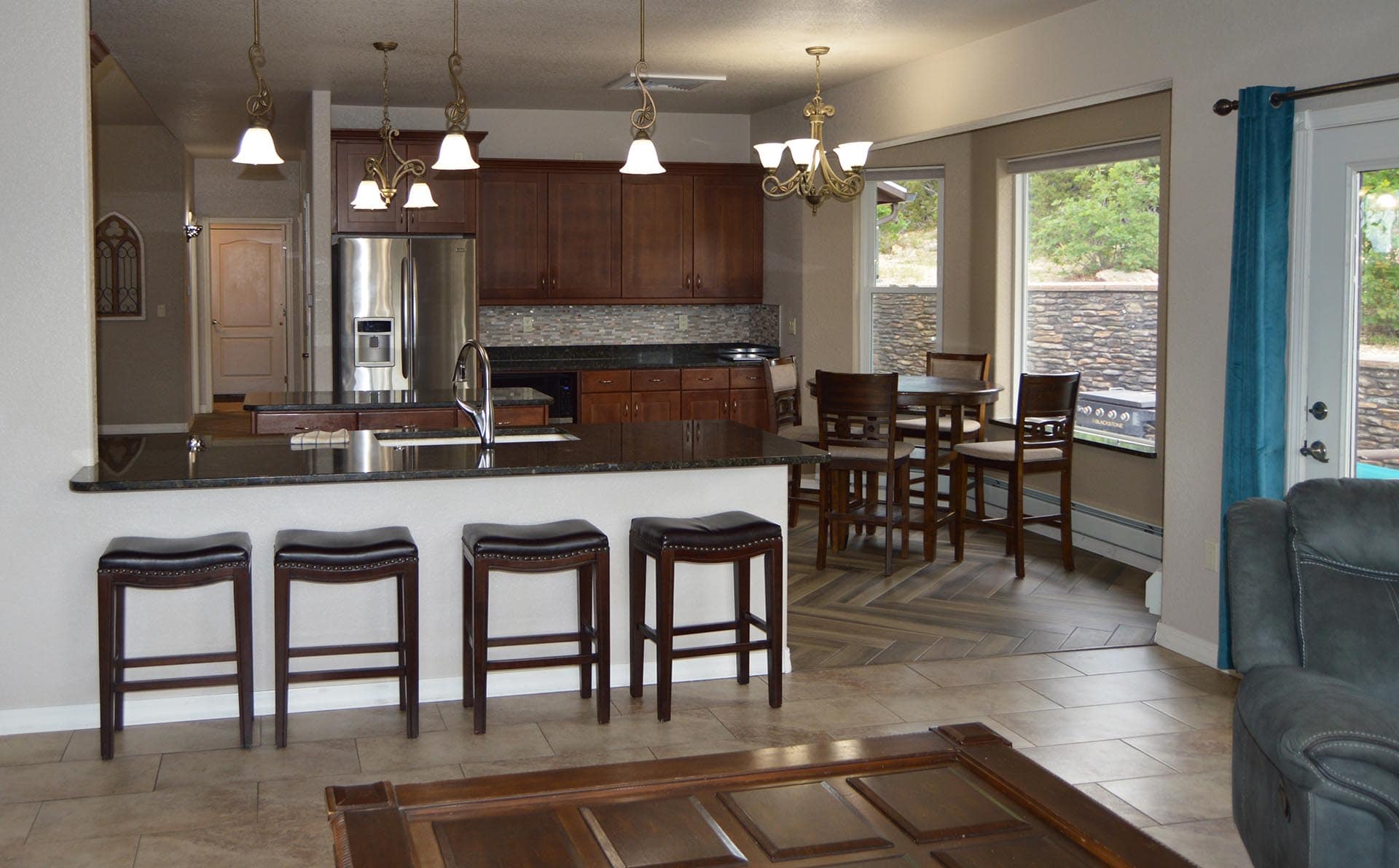 Open-plan kitchen with dark wood cabinets, stainless steel refrigerator, granite island, and four bar stools facing a dining area.