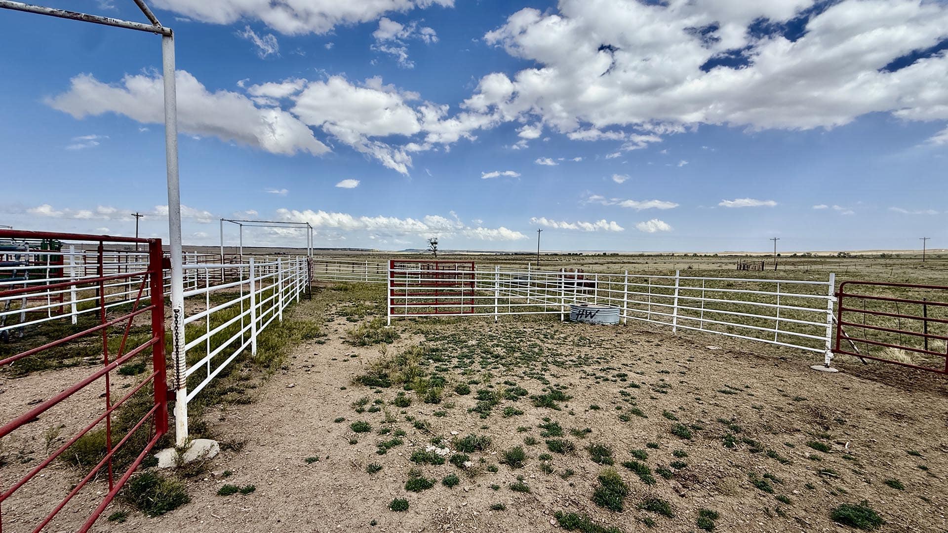 Wide rural cattle corral with red and white metal fencing under a blue sky with scattered clouds; dry, sparse ground and open plains in the distance.
