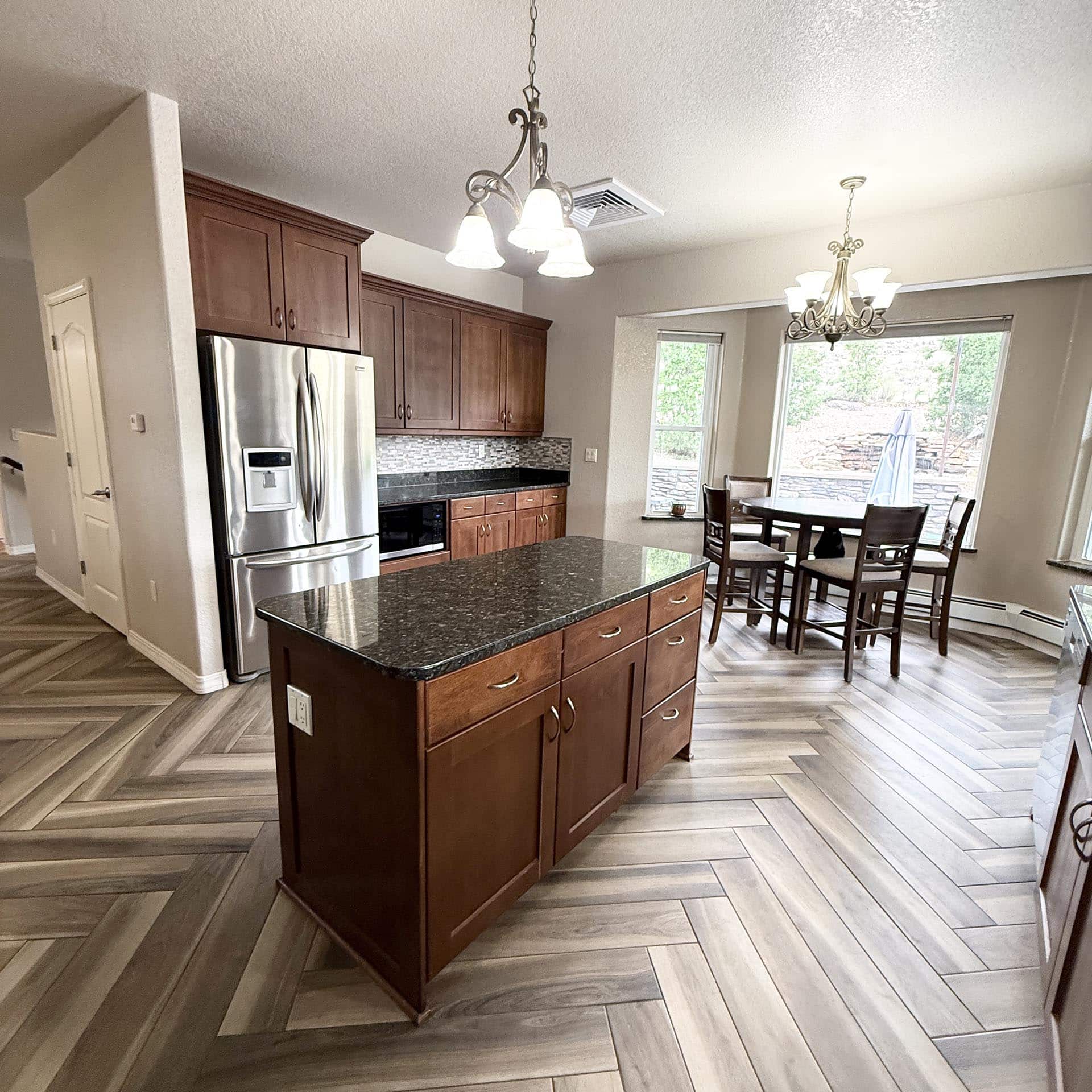 Kitchen with dark wood cabinets, stainless refrigerator, and granite island; hanging chandeliers illuminate a dining nook by large windows.