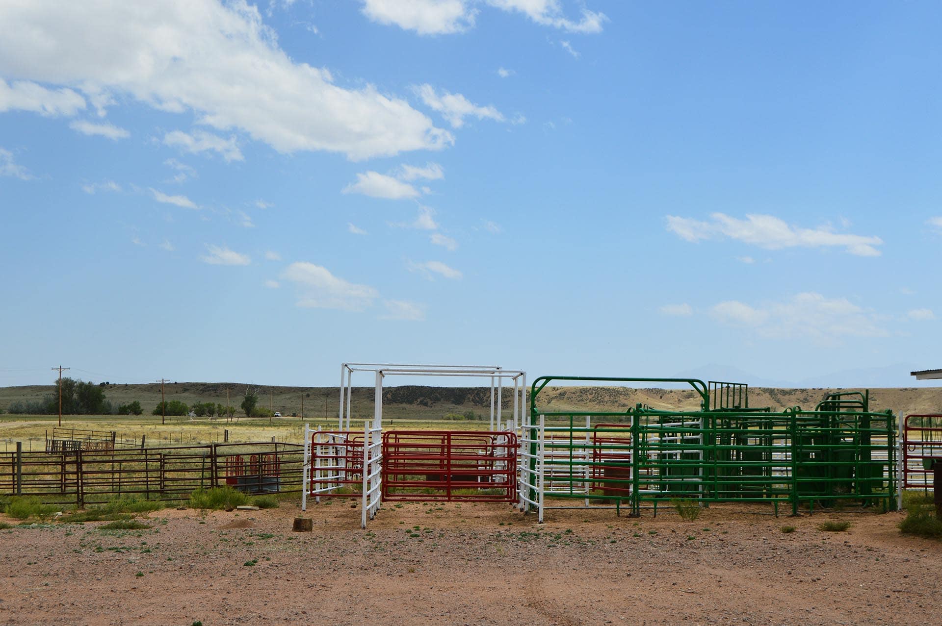 Open corral yard with stacked red, white, and green metal gate panels under a blue sky with scattered clouds.