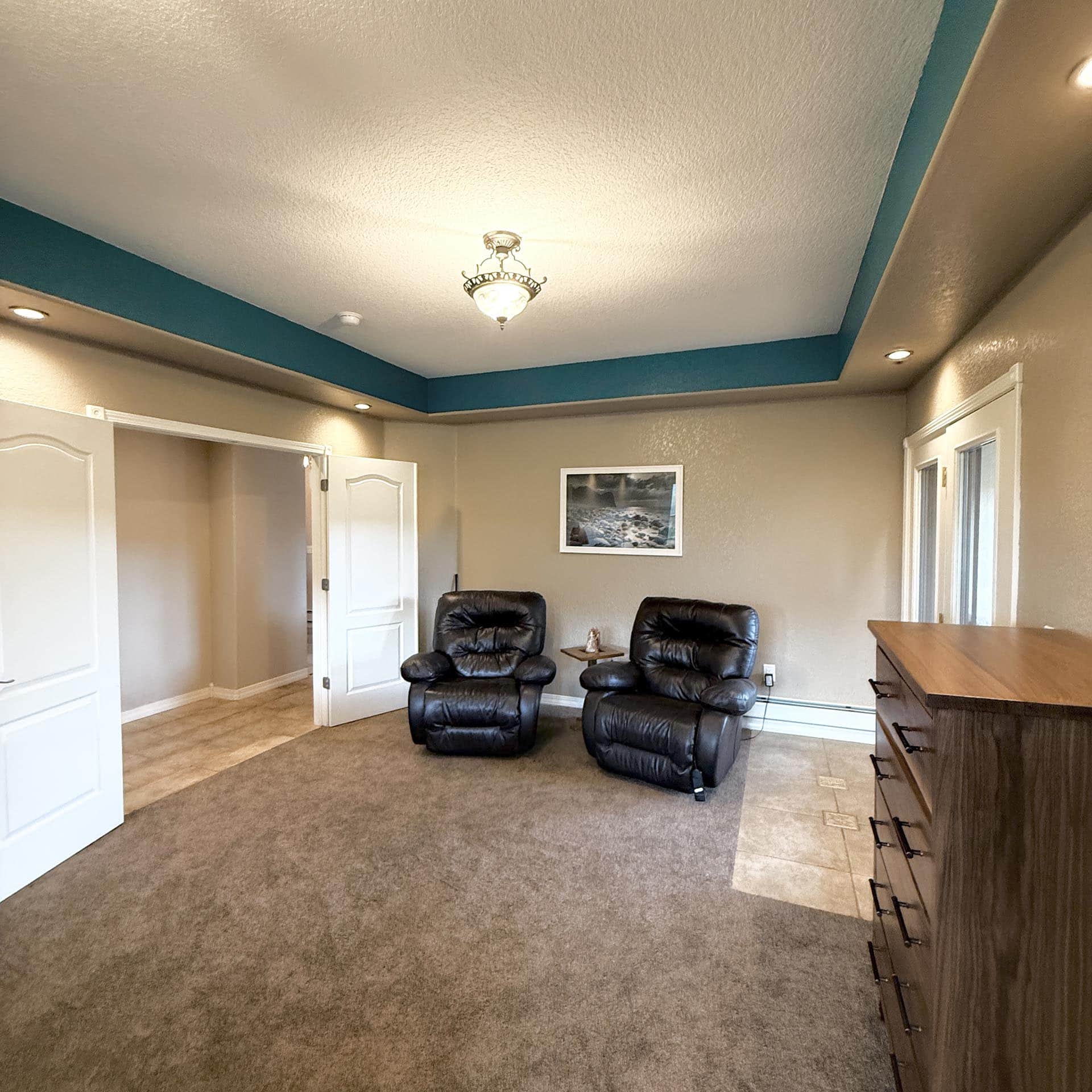 Living room or office seating area with two black leather recliners and a small table, teal trim along the ceiling, beige walls, and cabinets on the left/right edge of the image.