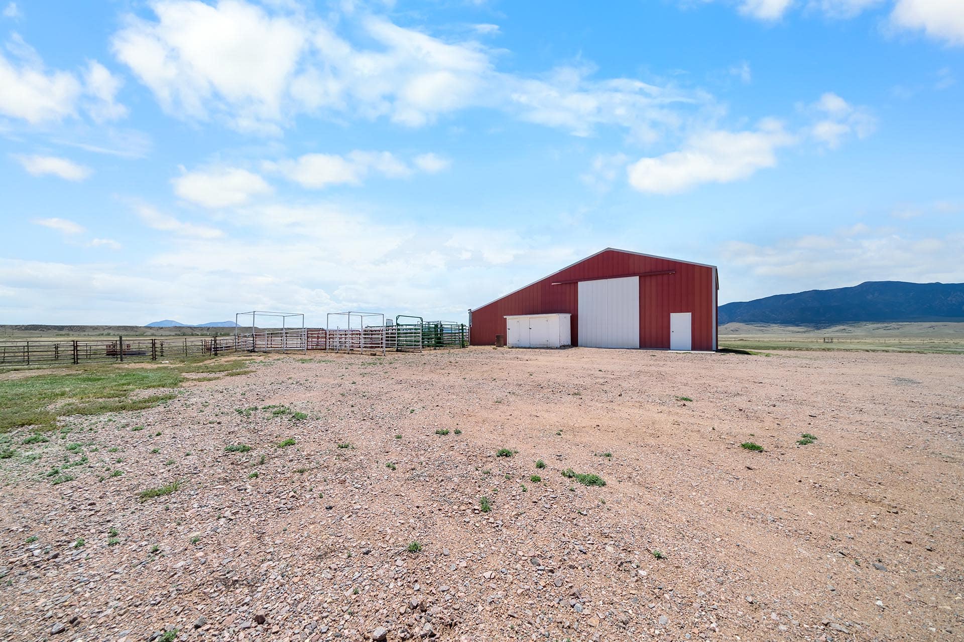 Red barn with white doors sits on a gravel yard under a bright blue sky and distant mountains.