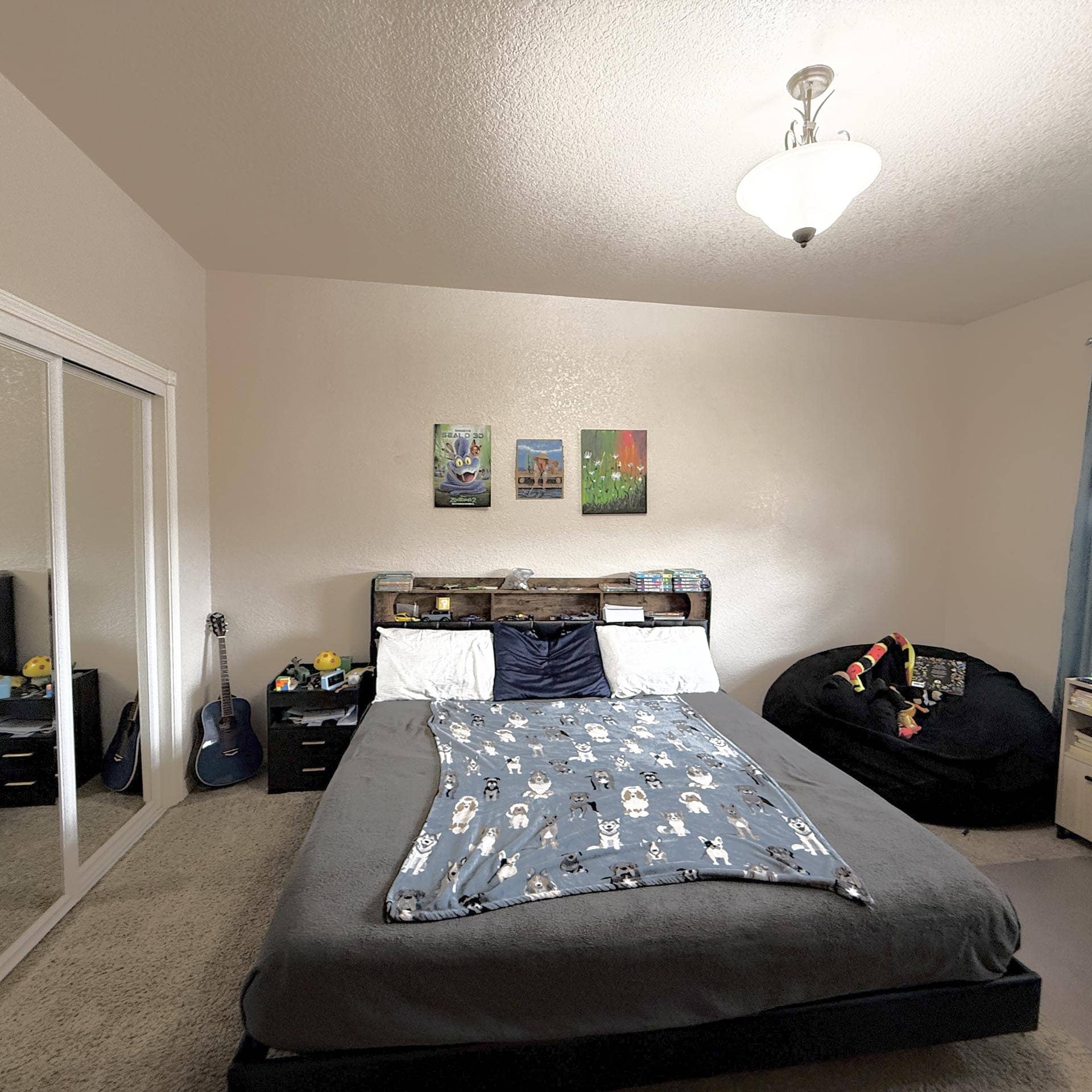 A bedroom with a low platform bed covered by a blue animal-print comforter, a guitar and small desk to the left, and framed pictures on the wall above the bed.