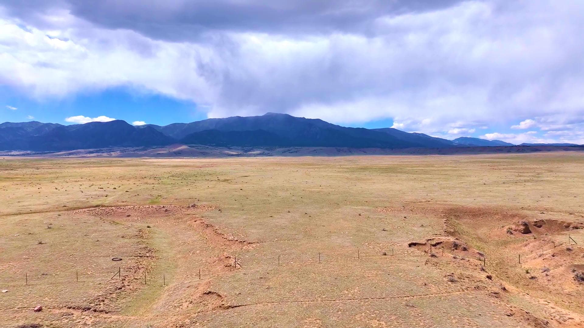 Wide arid plain with distant mountains under a cloudy sky and approaching rain in the distance.