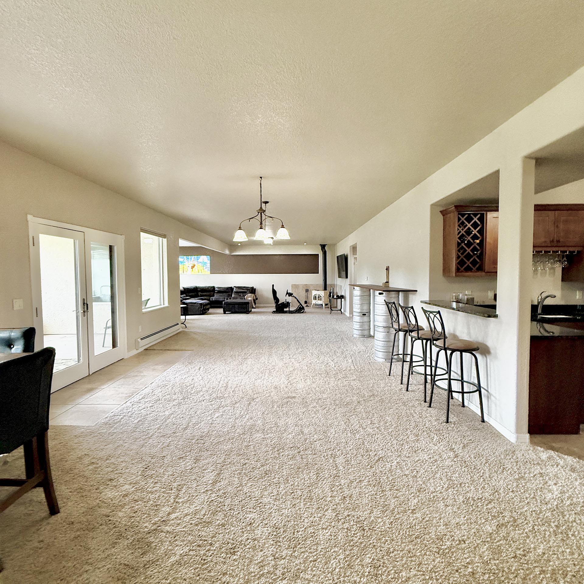 Open-plan living space with a kitchen bar and six metal stools along a counter, beige walls, and a seating area in the background.