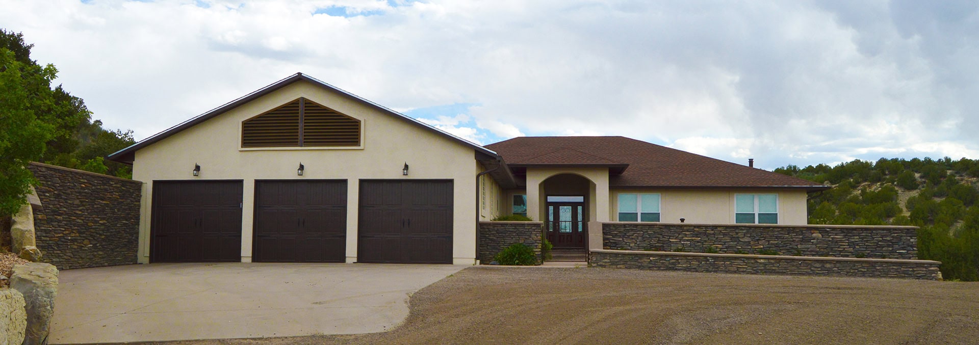 Three-car garage on a single-story beige house with stone accents and a front entry under a small arch, set on a hillside.