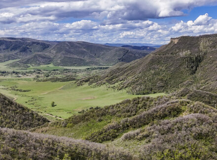 Panoramic view of a green valley surrounded by forested hills under a cloudy blue sky.