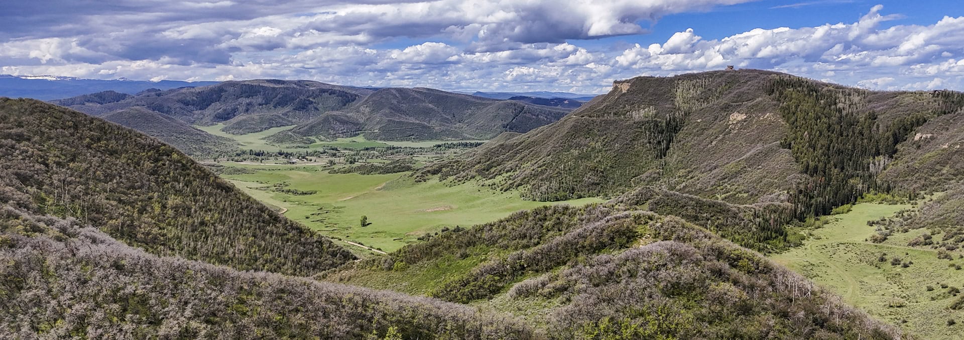 Panoramic view of a green valley surrounded by forested hills under a cloudy blue sky.