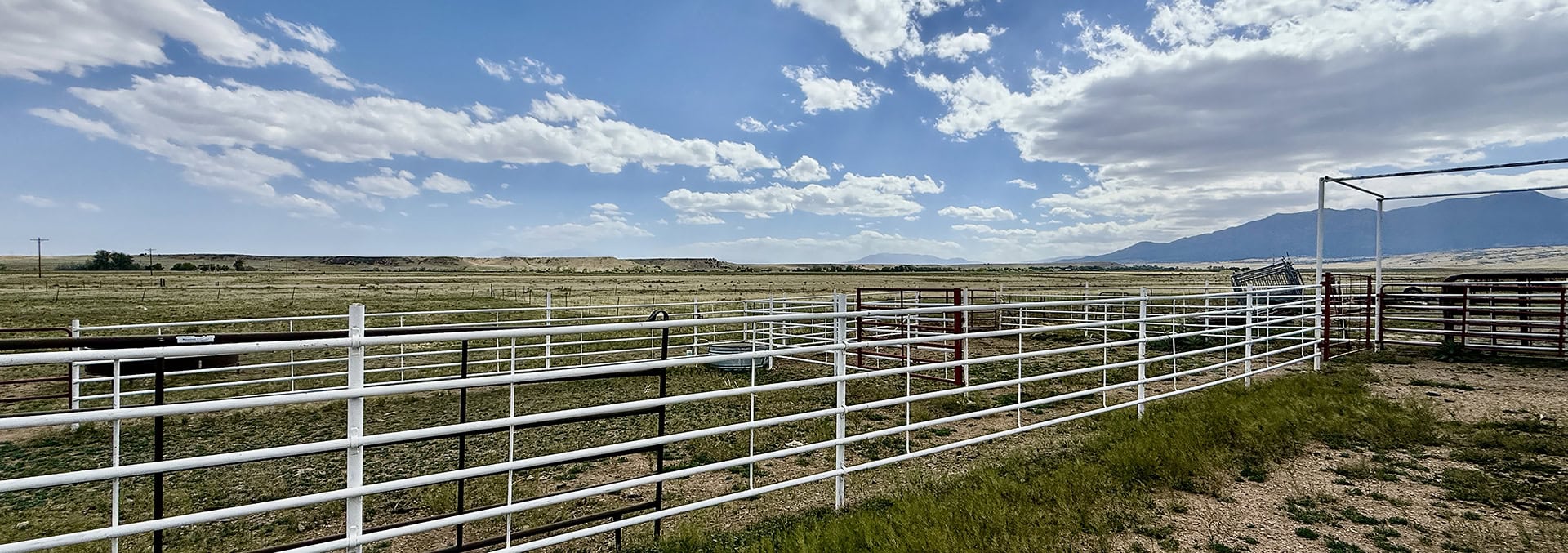 Wide view of a rural ranch with white metal fences, open fields, and distant mountains under a blue, cloud-speckled sky.