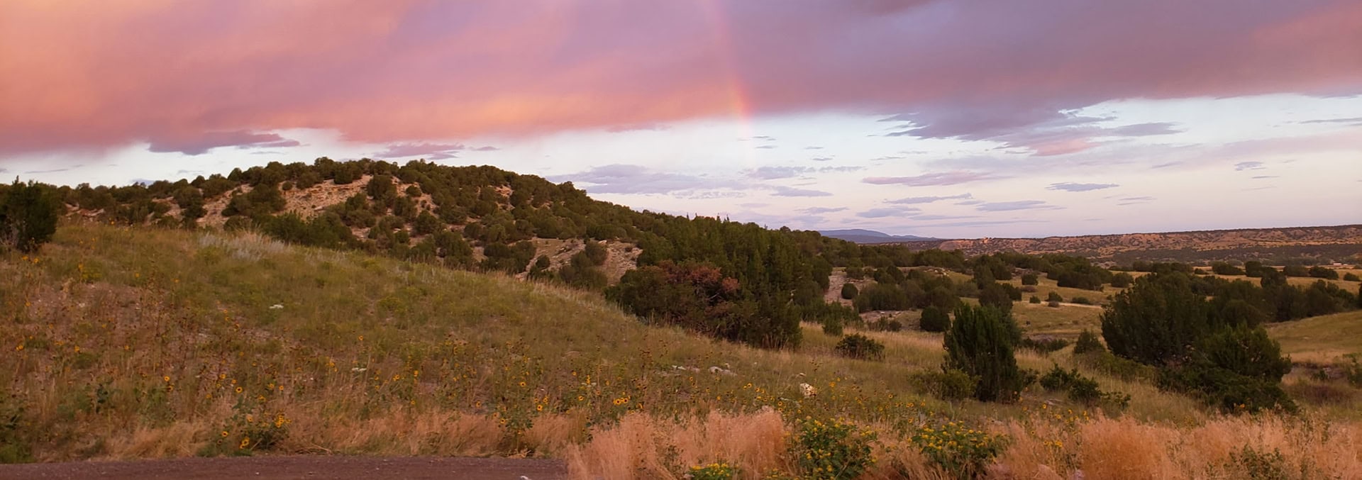 Sunset sky over rolling hills with a faint rainbow and wildflowers in the foreground, creating a peaceful prairie scene.