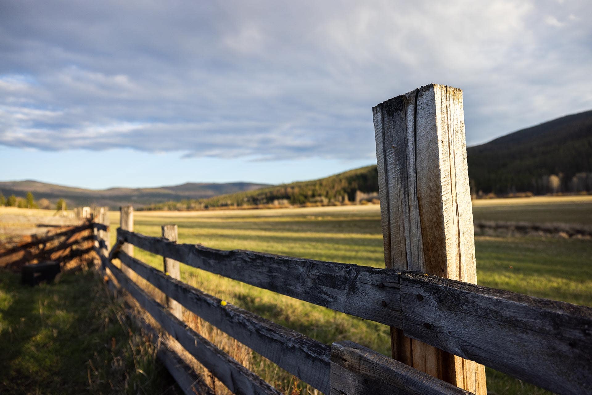 Wooden split-rail fence in a rural field with rolling hills under a cloudy sky.