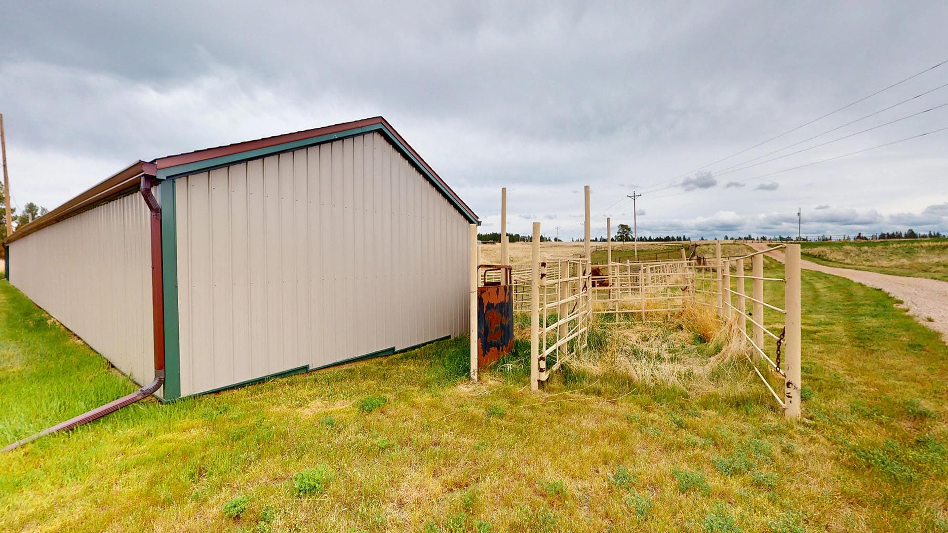 Long white corrugated-metal barn with red trim beside a beige fence in a grassy rural field under a cloudy sky.