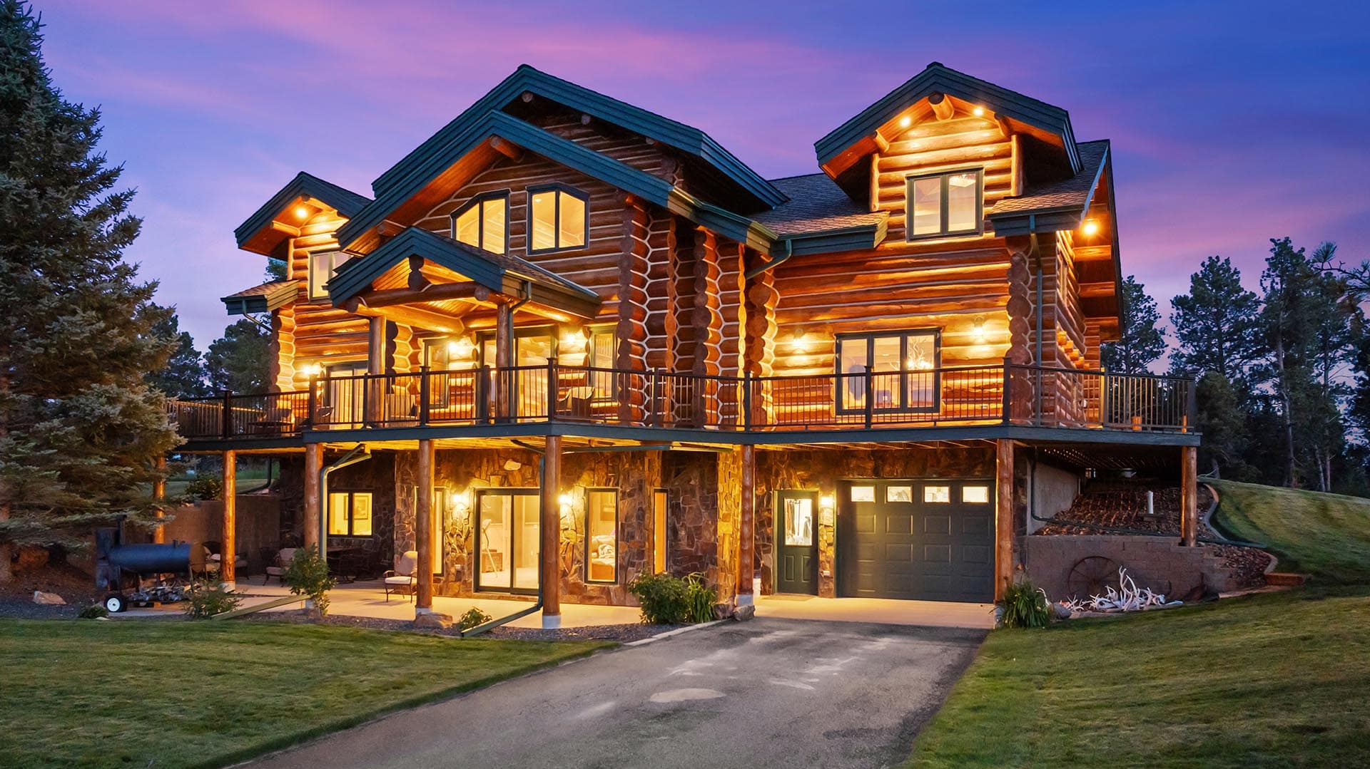Two-story log cabin with a large wraparound deck, warm interior lights, and stone foundation, set against a twilight sky and trees.