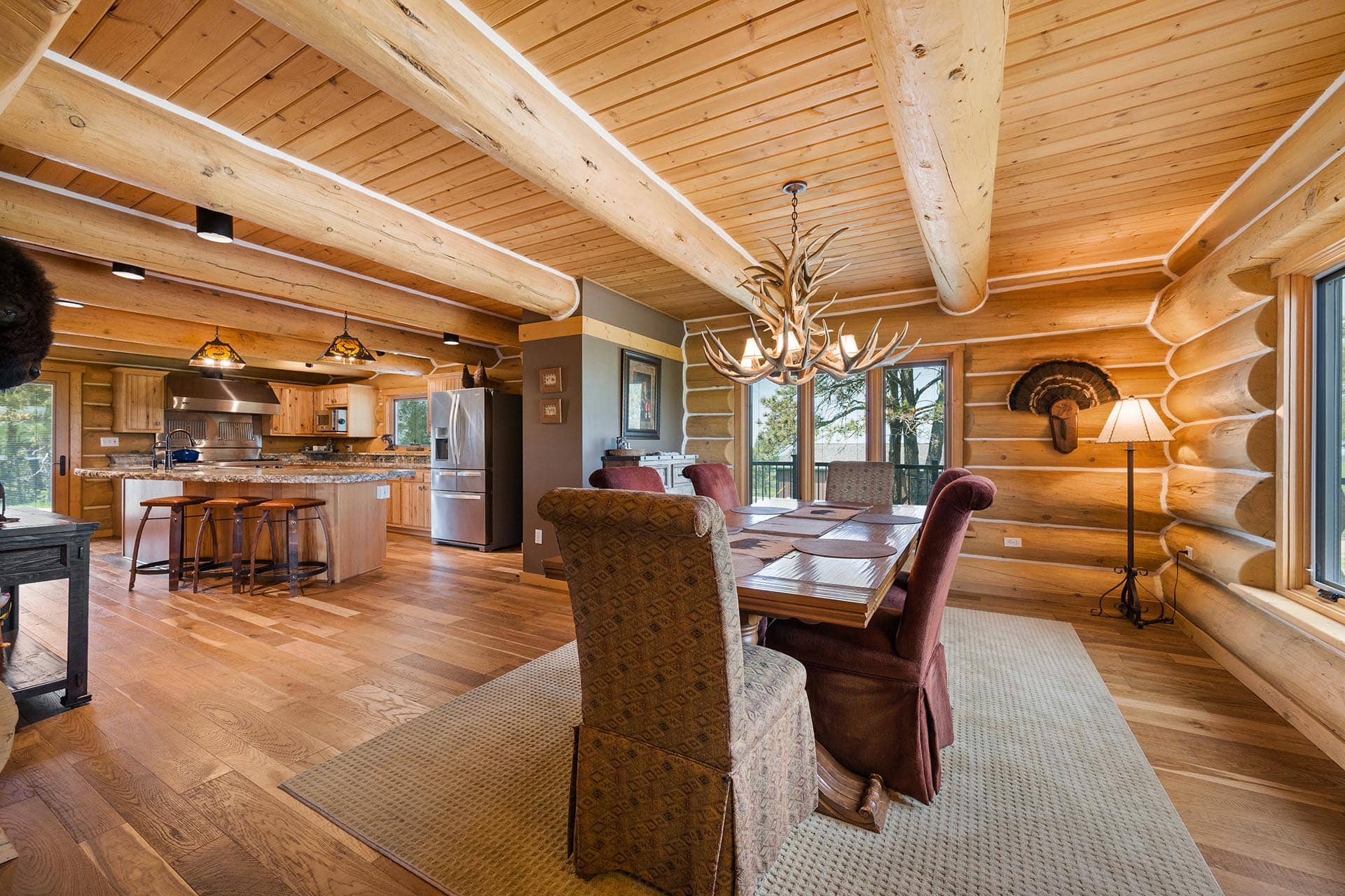 Log cabin interior with exposed ceiling beams, a granite island, and a dining table under an antler chandelier in a rustic kitchen/dining area.