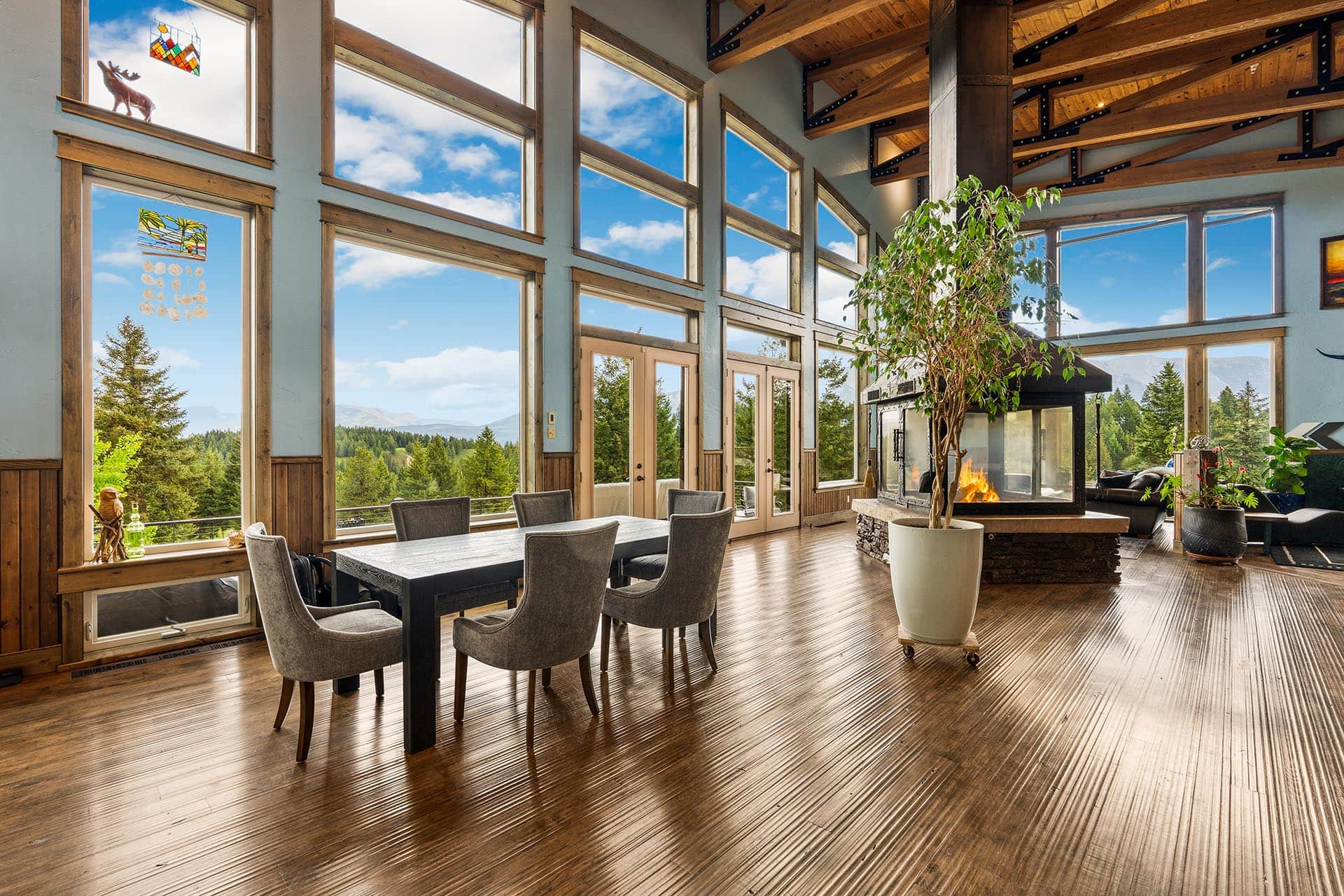 Bright lodge-style living room with large windows, a dining table and grey chairs, a fireplace, and a forested mountain view outside.