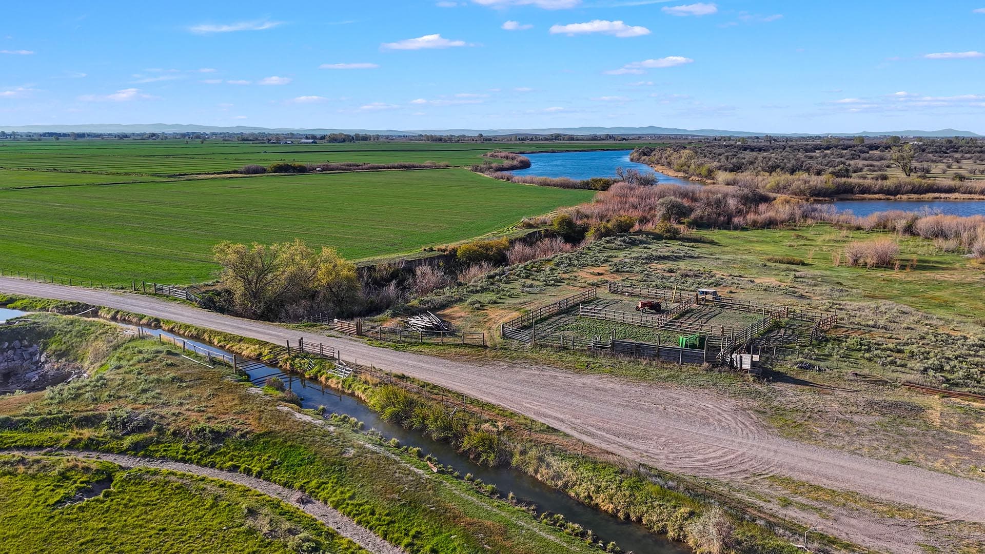 Aerial view of green fields, a winding river, and fenced pastures with farm equipment under a blue sky.