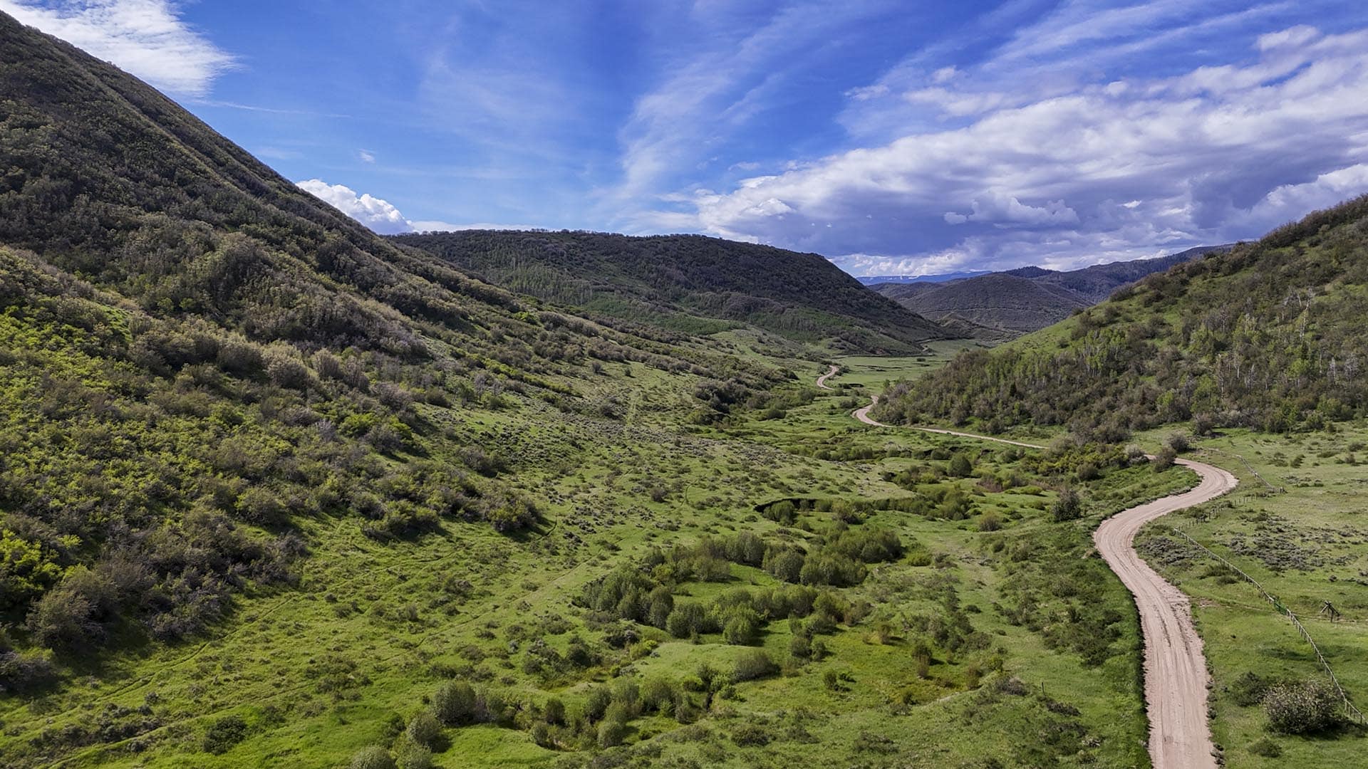 Wide valley with grassy fields and rolling hills, a winding dirt road cutting through the center under a blue sky.