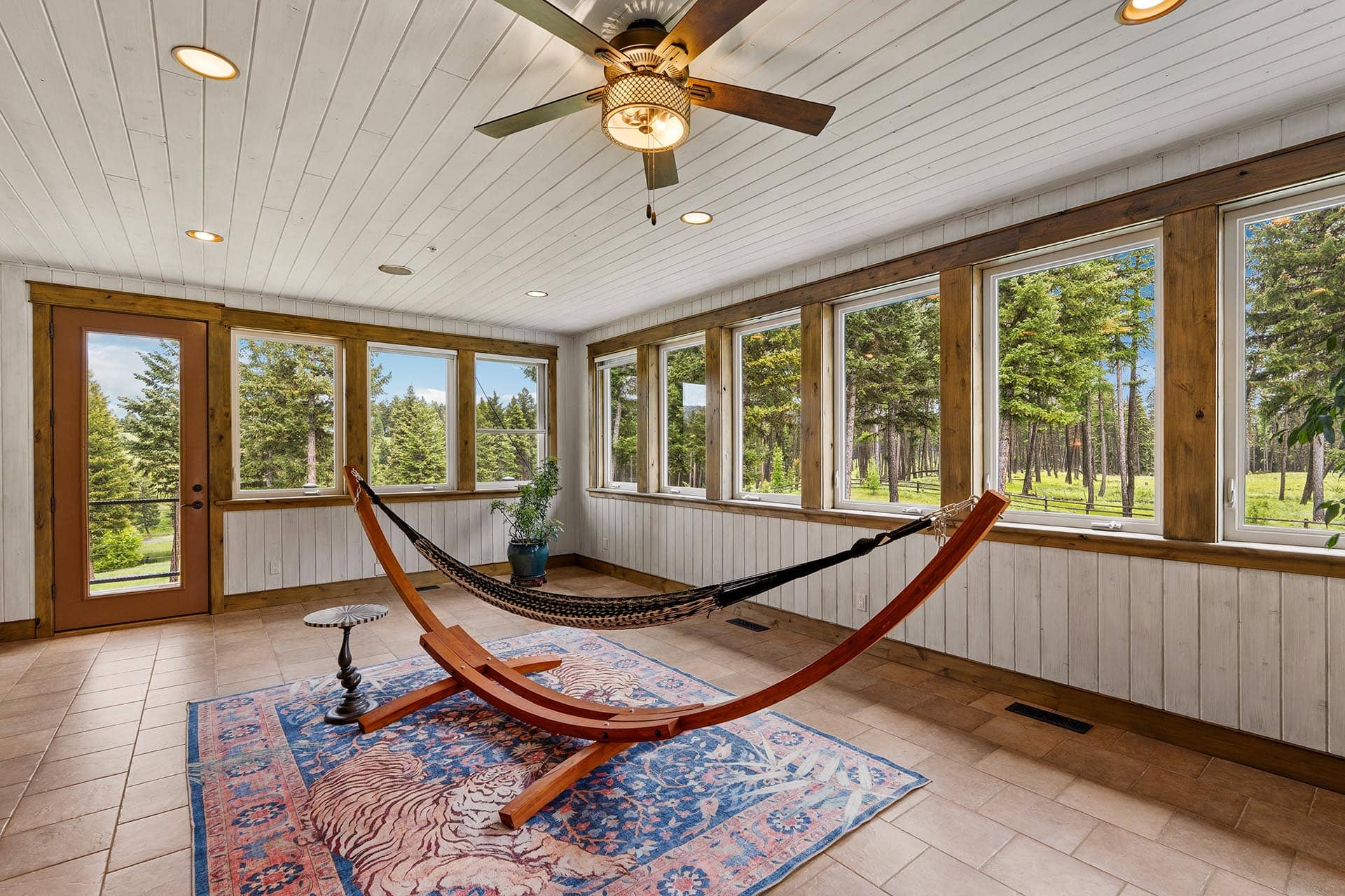 Sunlit sunroom with a wooden hammock, blue patterned rug, and large windows showing a forest outside.