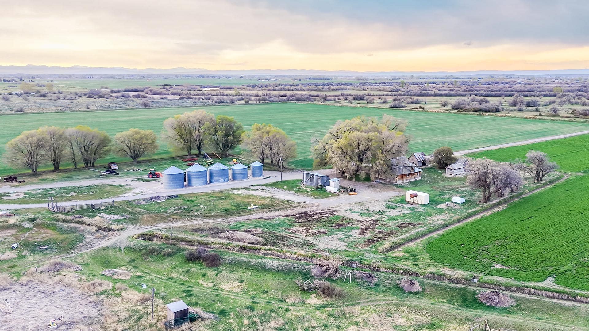 A farmyard with several metal grain silos, a red tractor, sheds, and surrounding trees amid green fields under a cloudy sky.