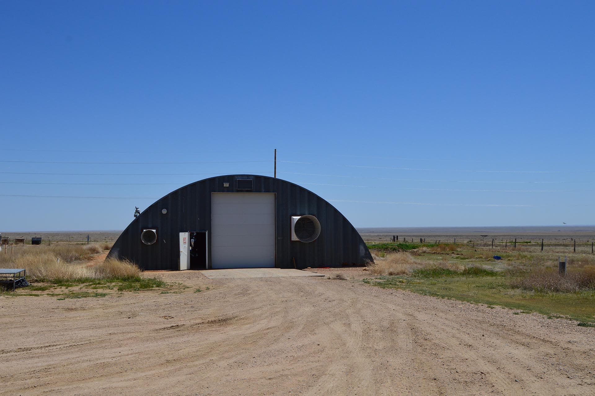 Curved metal barn in a dry, flat desert landscape under a clear blue sky, with a white door open at the front and large circular vents on either side.