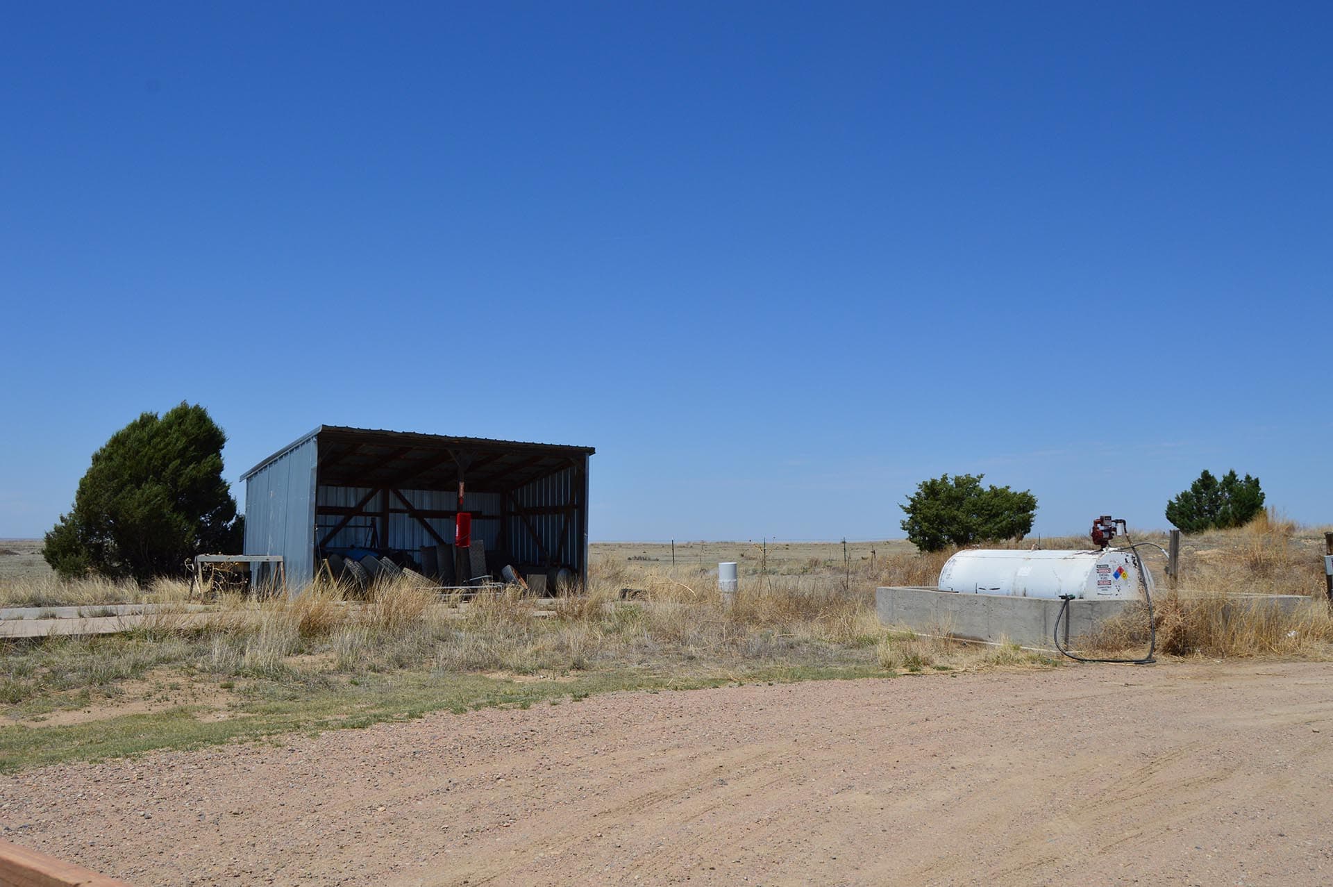 Open rural scene with a blue metal shed left, dry grass, and a white tanker trailer on the right under a clear blue sky.