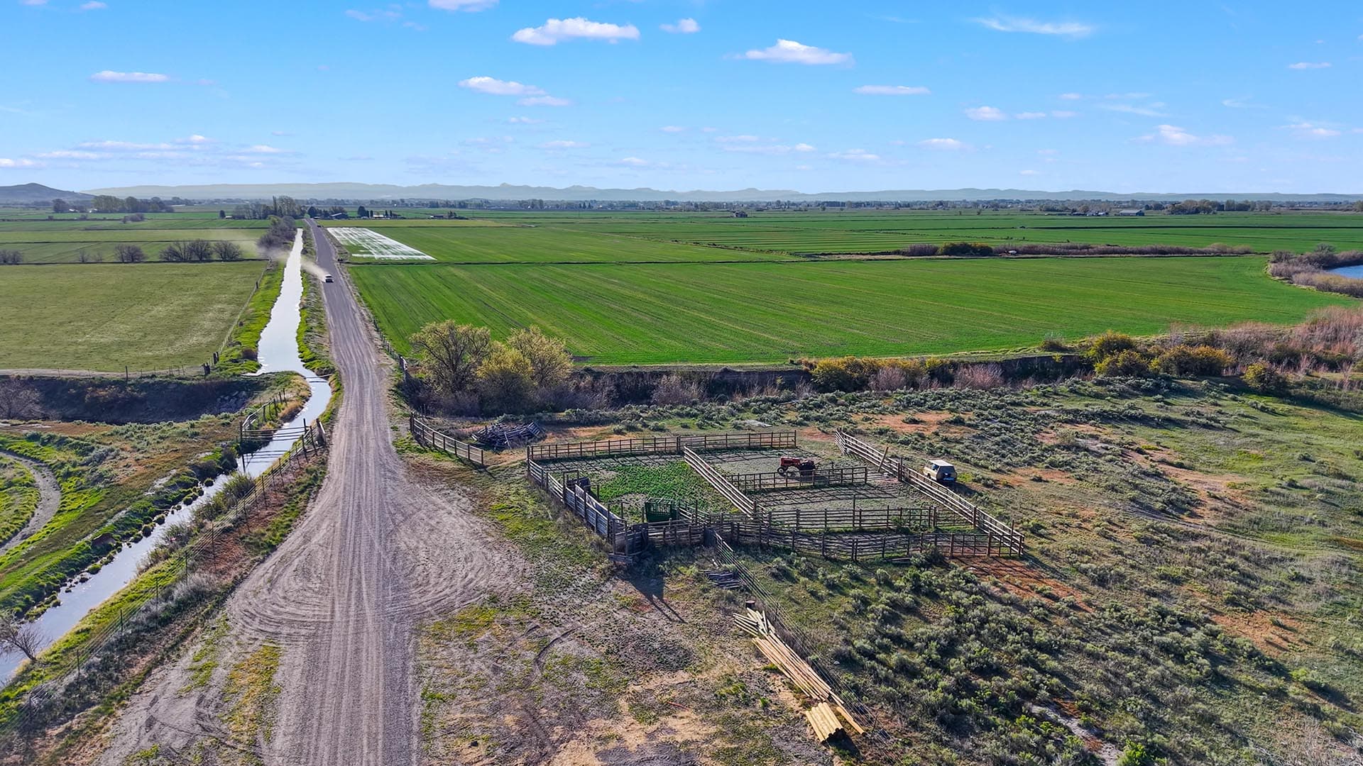 Aerial view of a rural farm: green fields, a dirt road, a canal, and fenced animal pens beneath a blue sky with distant hills.