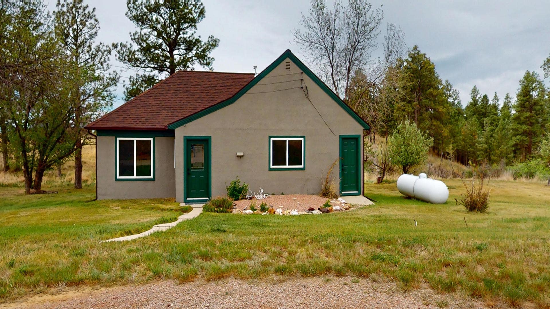 Small beige house with green trim and doors, red roof, in a grassy yard with trees and a white propane tank to the right.