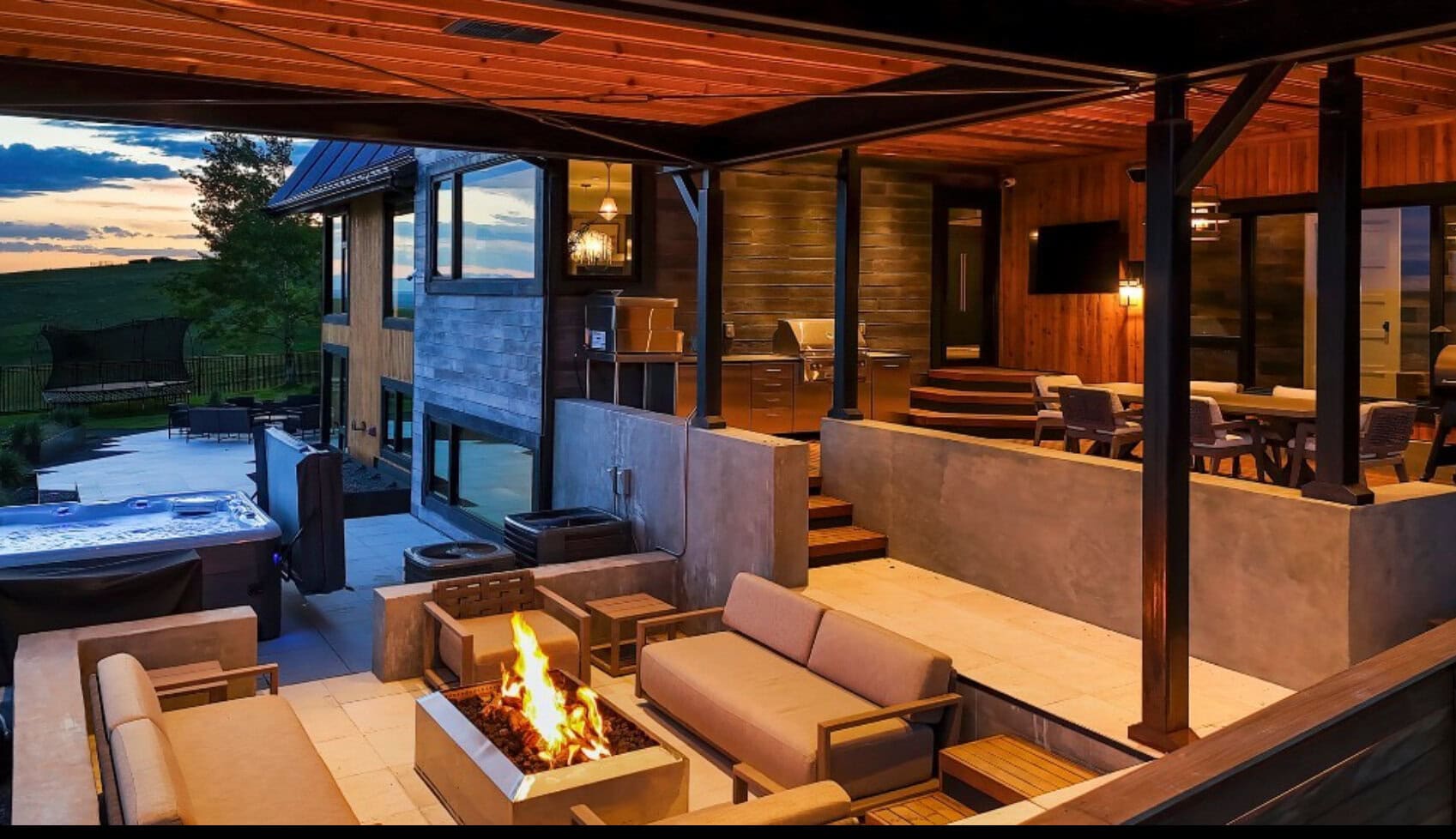 Modern outdoor living space with a fire pit, beige sofas, and a covered patio overlooking a yard and hot tub area at dusk.