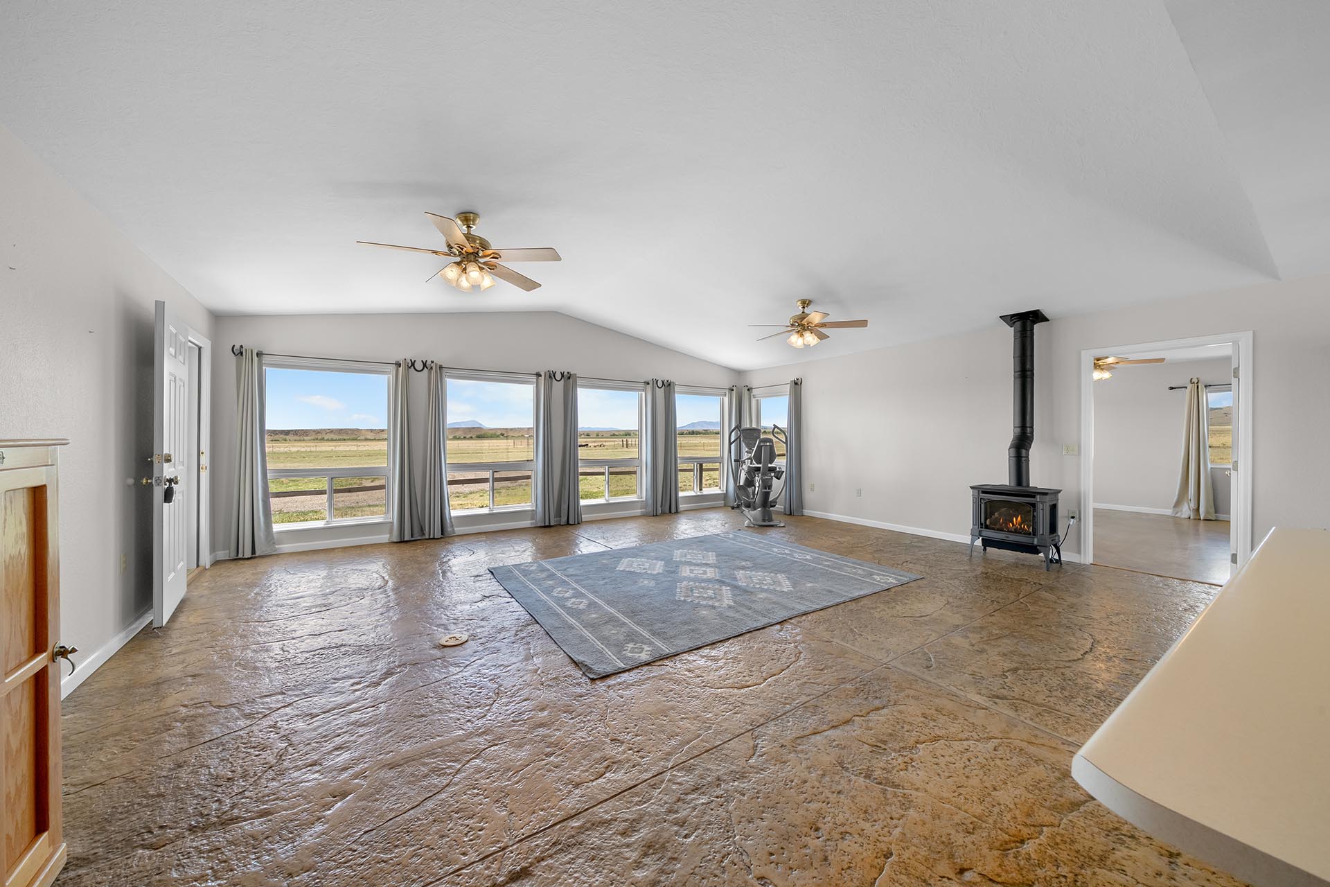 Bright living room with large windows showing countryside; two ceiling fans, wood stove, and exercise bike along the wall.