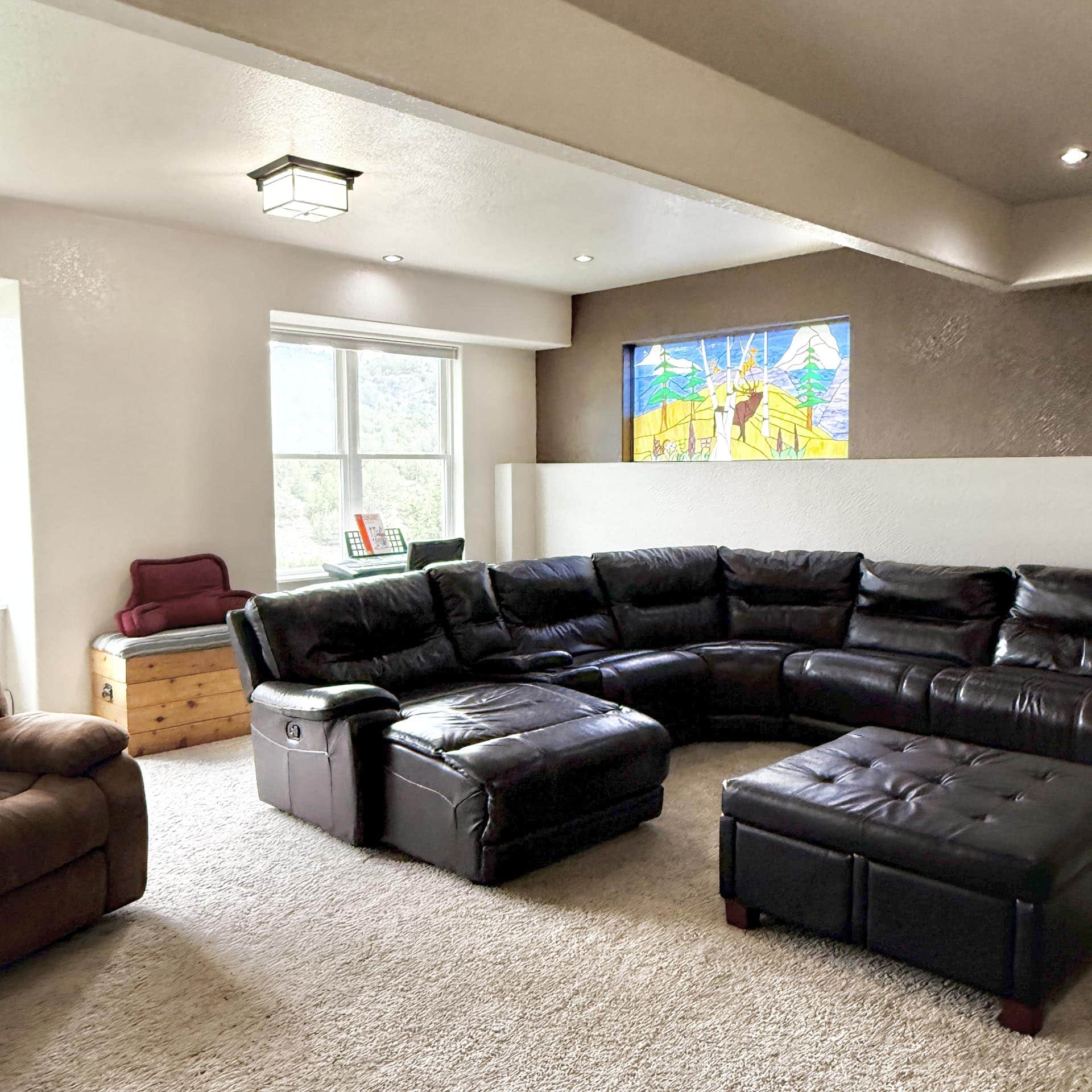 A spacious living room with a large dark leather sectional sofa, beige carpet, and a colorful stained-glass window on the wall.