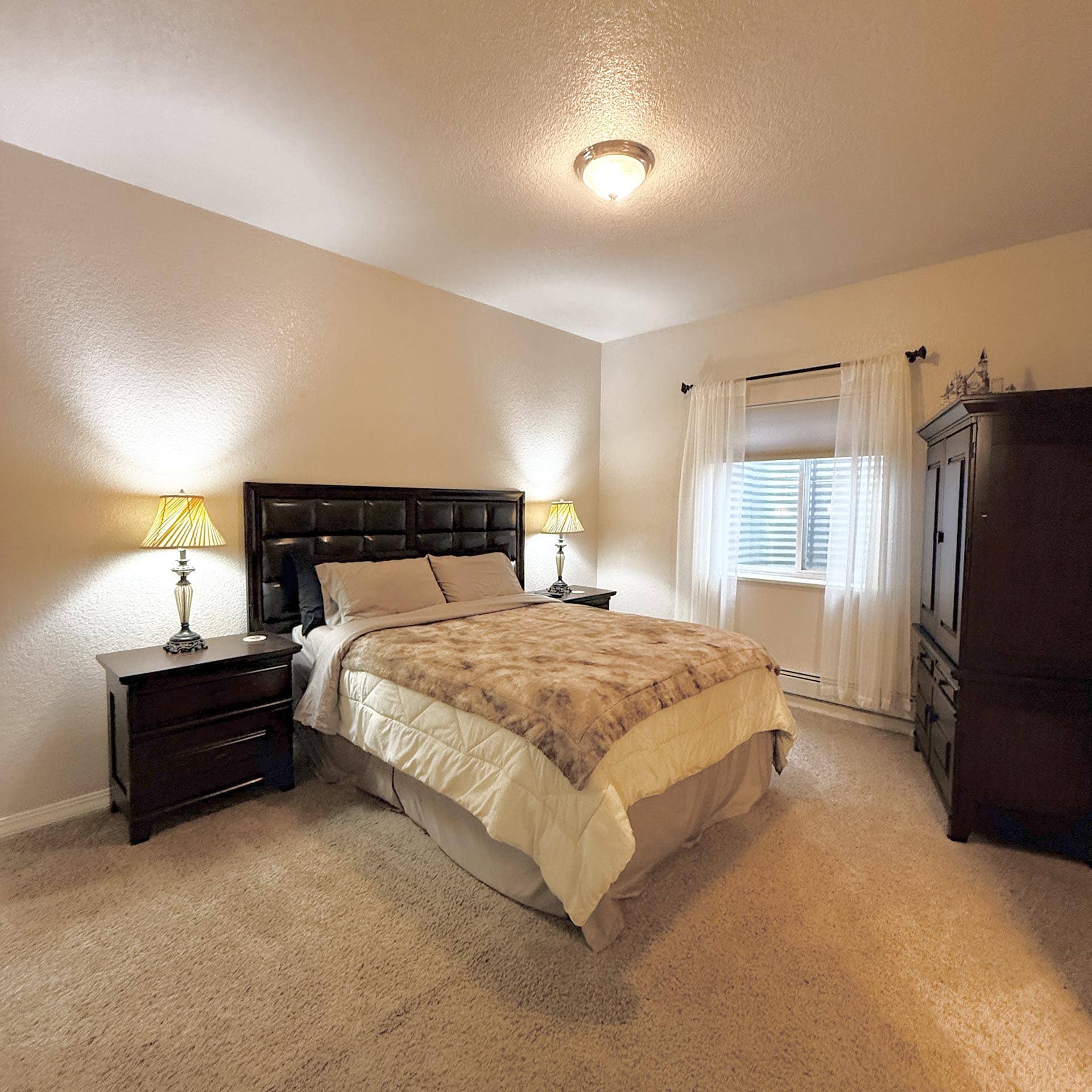 Bedroom featuring a dark wood queen bed with beige bedding, two matching nightstands with lamps, and a dresser by a window with sheer curtains.
