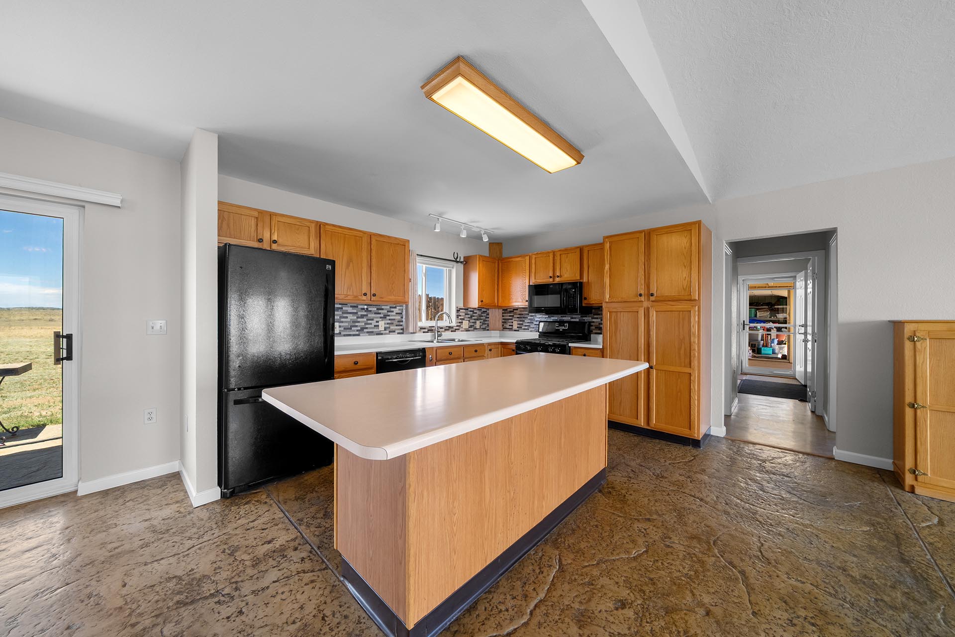 Open-concept kitchen with an island, oak cabinets, a black refrigerator, and a tiled floor/mapled ceiling light fixture in the center of the room.