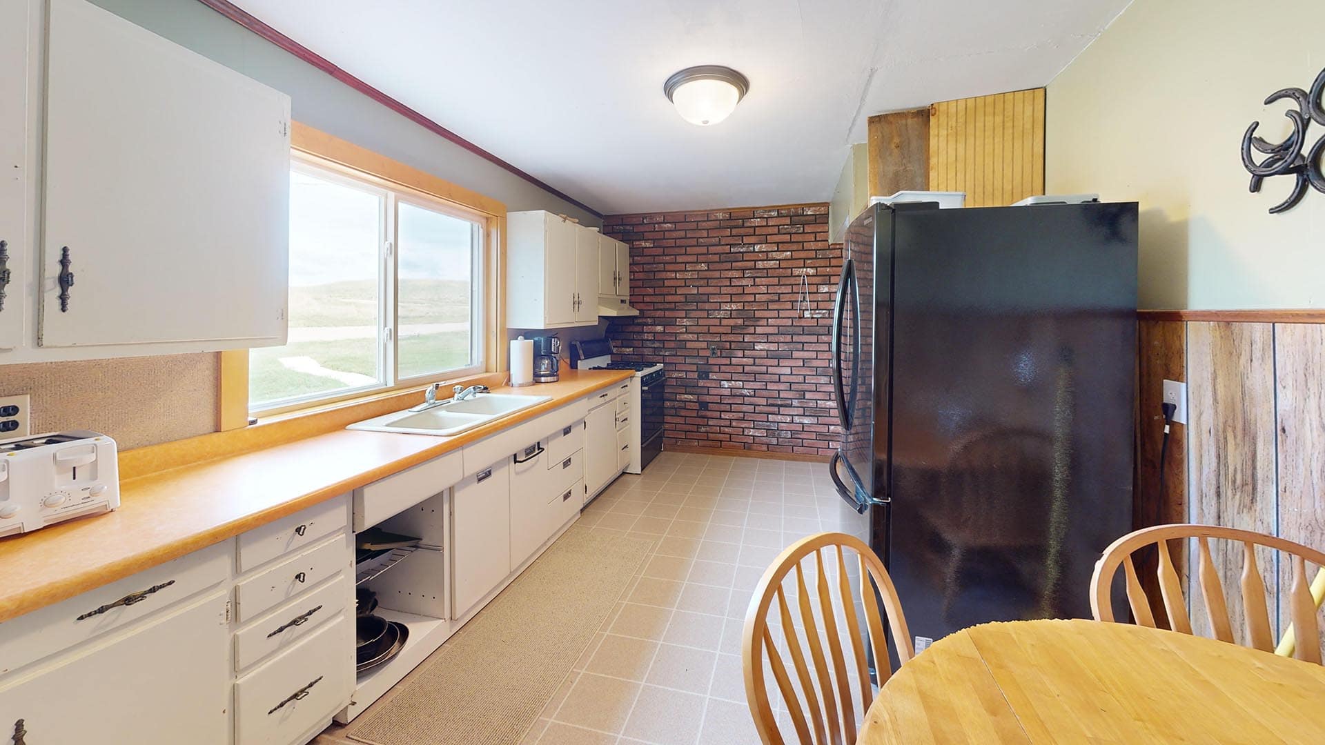 Bright kitchen with a brick accent wall at the far end, white cabinets, light wood countertops, and a large black refrigerator, with a round wooden dining table and chairs in the foreground.