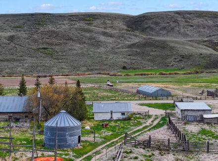 Rural farmstead with a metal silo, sheds, fences, and a winding road amid rolling brown hills behind.