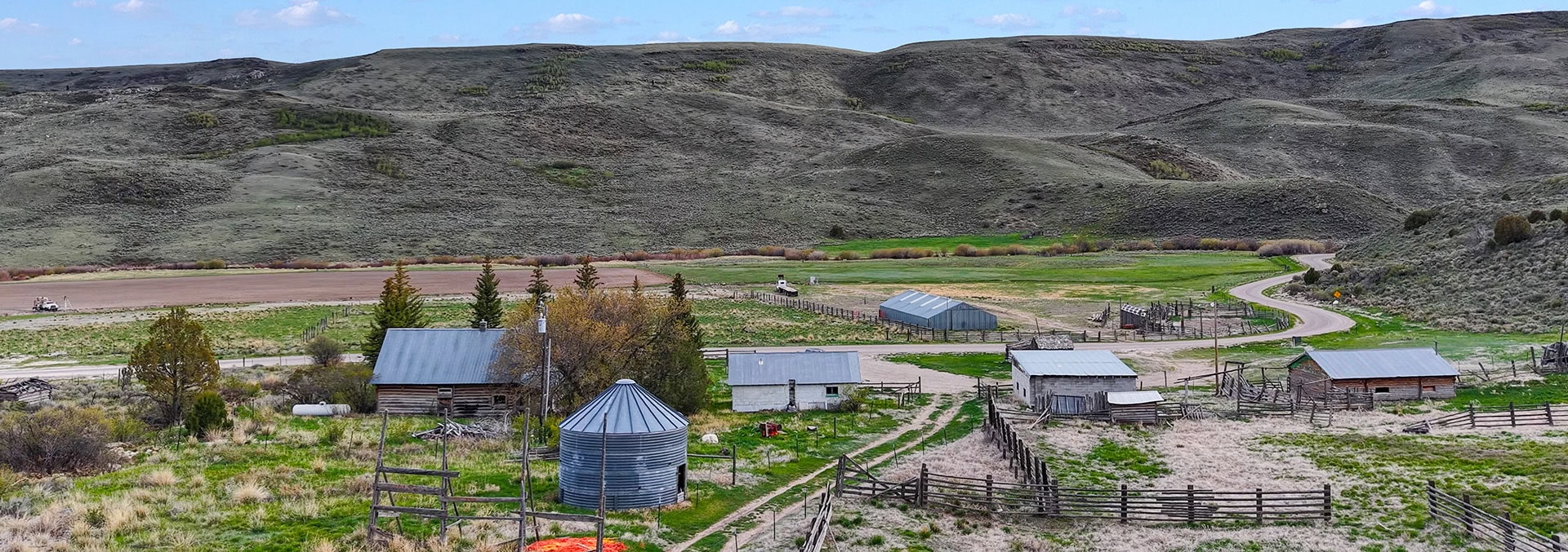 Rural farmstead with a metal silo, sheds, fences, and a winding road amid rolling brown hills behind.
