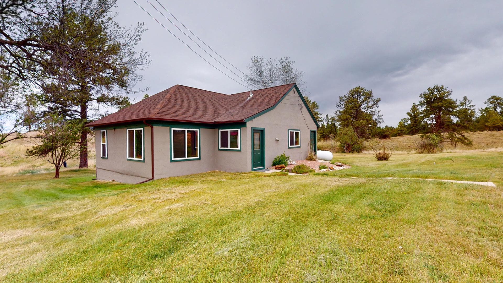 Single-story beige house with green trim and a brown roof in a wide grassy yard under an overcast sky, rural setting.