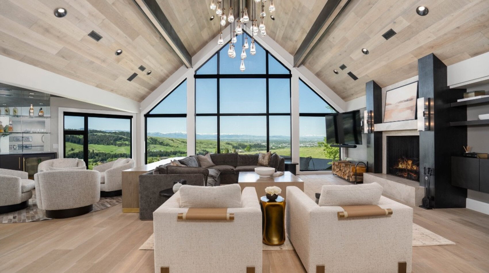 Luxurious living room with a cathedral wooden ceiling, oversized glass wall and panoramic countryside view beyond the sofa area.